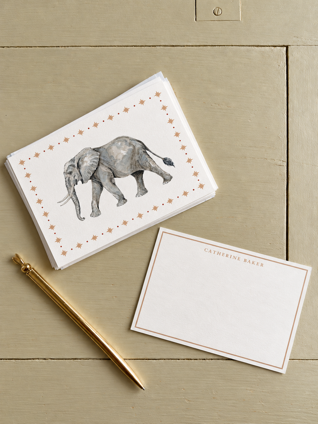 A stack of cards featuring a watercolor illustration of an elephant, surrounded by small star accents. Next to the cards is a gold pen and a blank stationery card with the name Catherine Baker printed at the top.