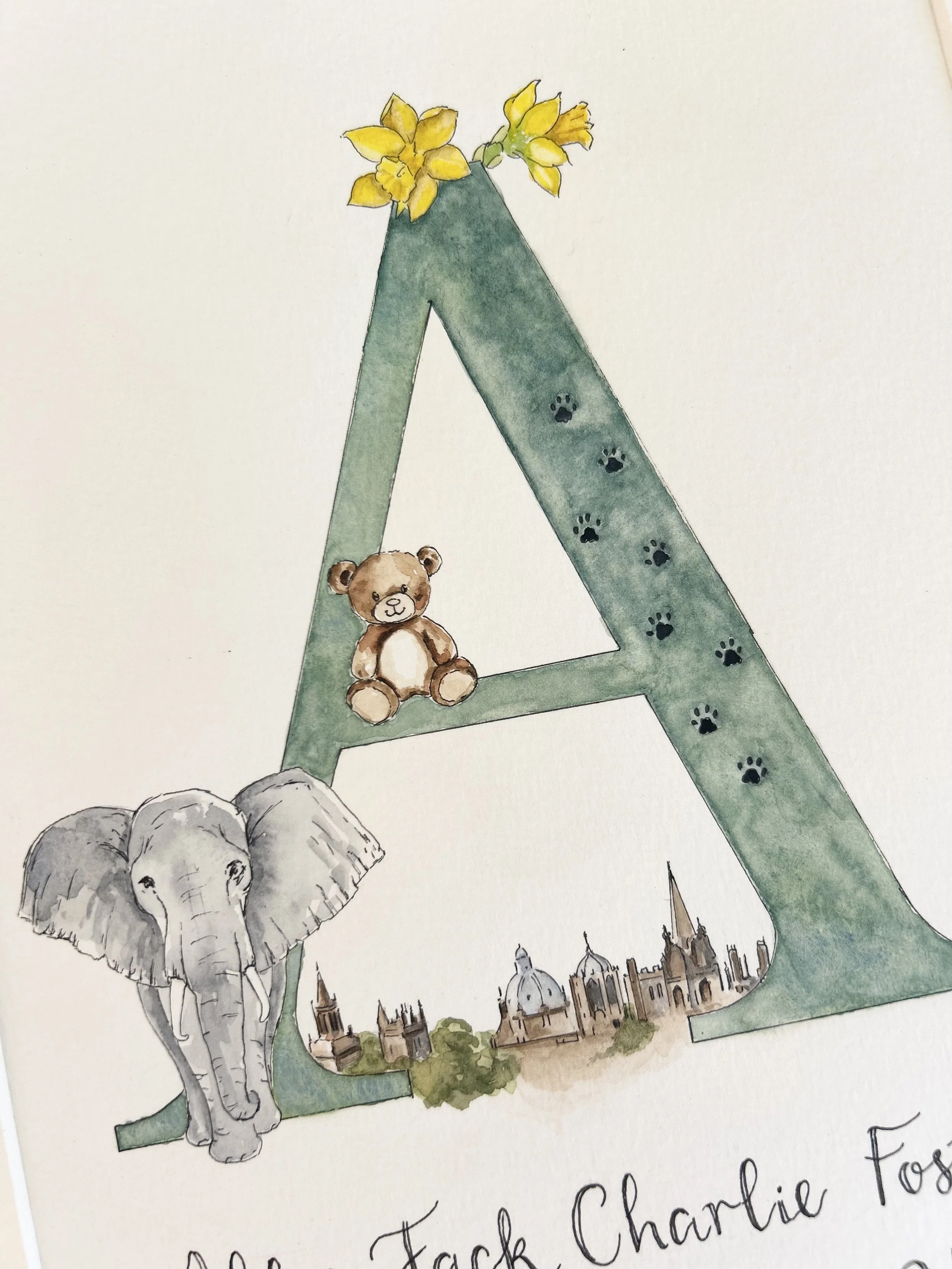 A watercolor illustration of the letter 'A' decorated with yellow flowers at the top, black paw prints along the side, a small teddy bear sitting inside the letter, an elephant leaning against it, and a cityscape with historical buildings at the bott