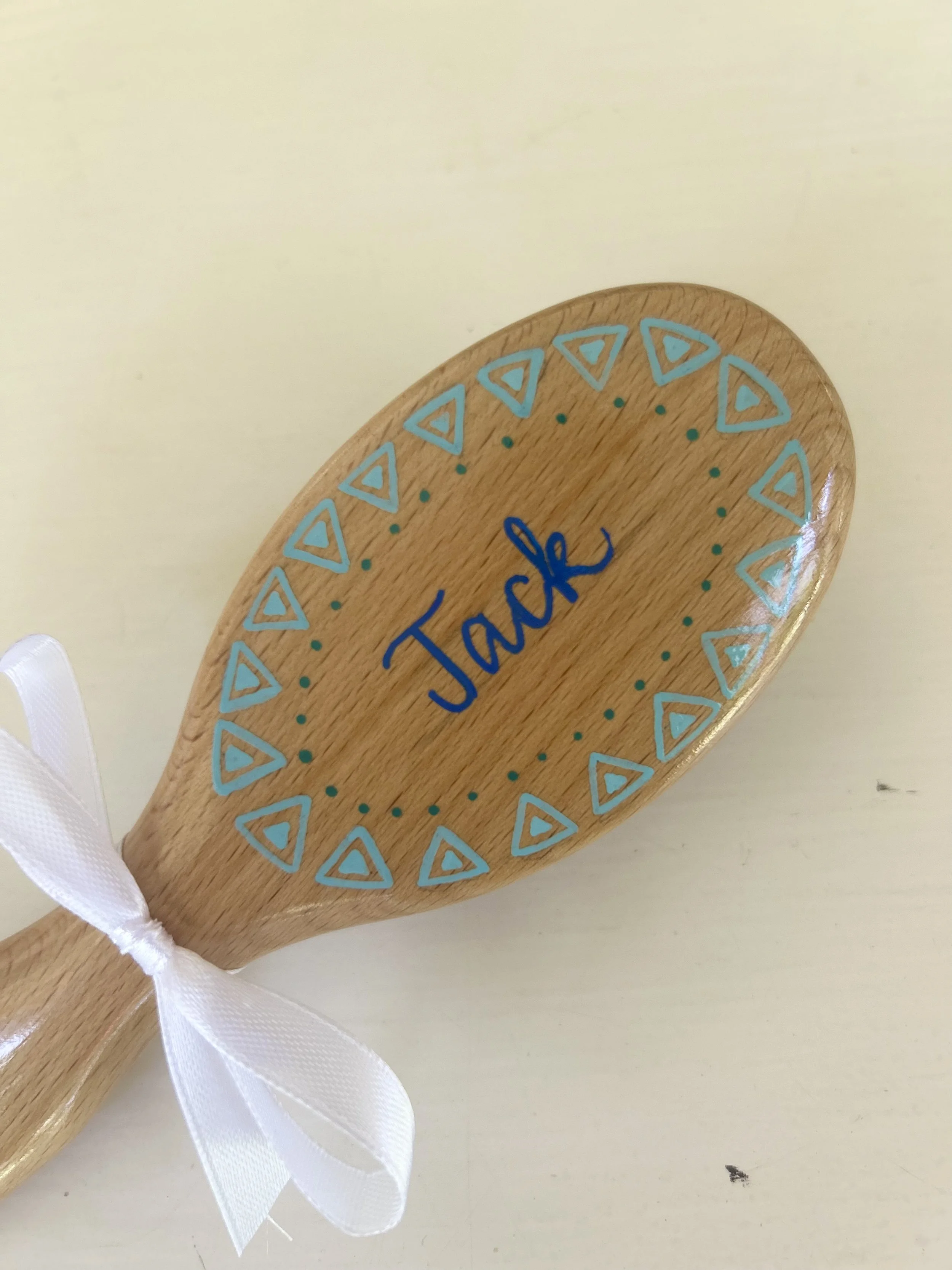 A wooden baby brush with the name 'Jack' written on it, tied with a white ribbon, placed on a white cloth on a wooden table. In the background, there is a bowl filled with white cotton balls, a tube of lotion, and a wicker basket filled with rolled-up towels.