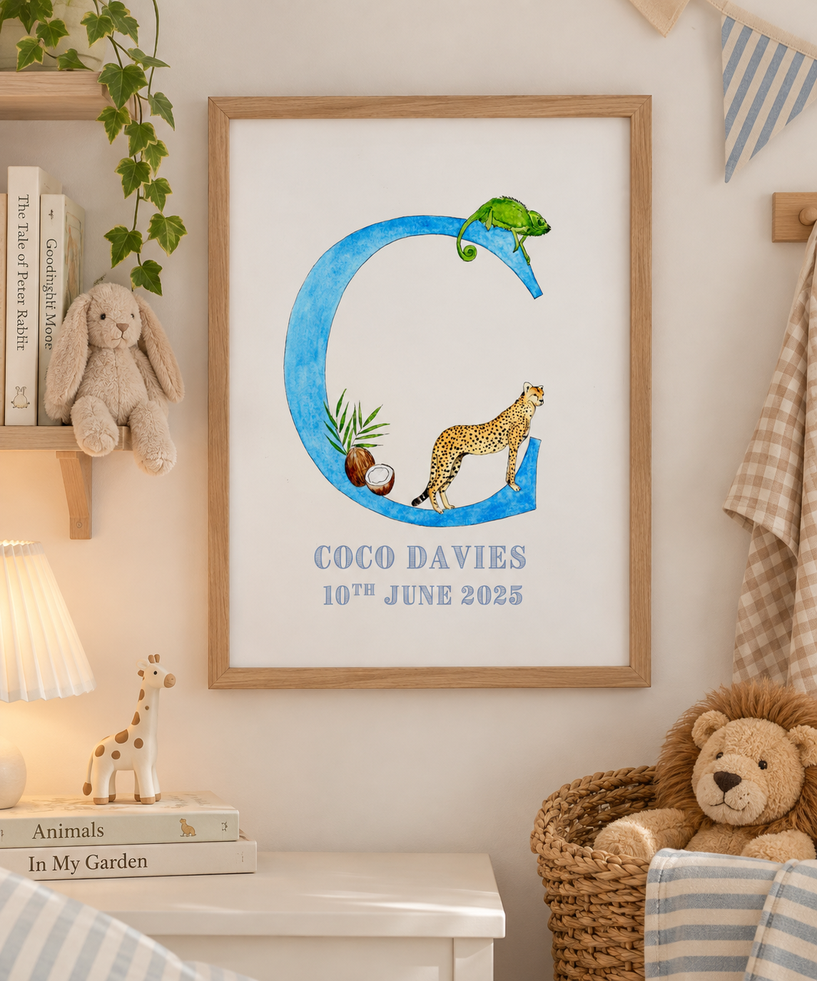 Children's room decorated with a framed artwork featuring a large blue letter C with a chameleon on top and a cheetah inside the letter, along with coconut and plant illustrations. The artwork displays the name Coco Davies and the date June 10, 2025. The room has plush toys, a small lamp, books, and striped and checked fabric decor.