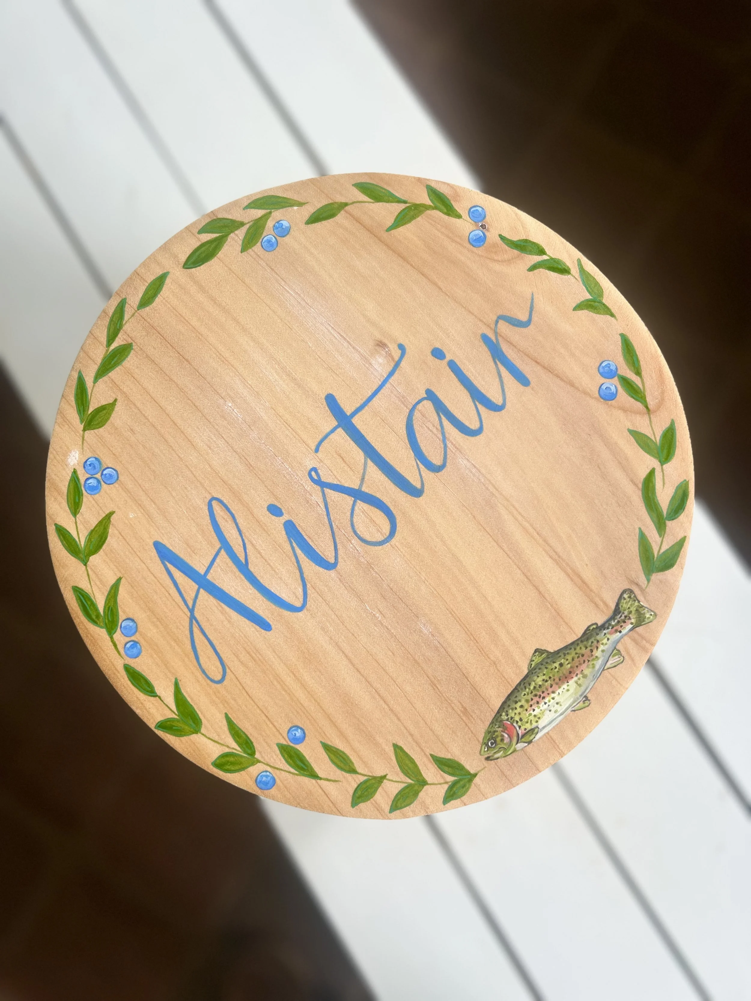 Close-up of a decorated round wooden sign with a border of green leaves and blue berries, featuring the word 'fish' and a colorful fish illustration.