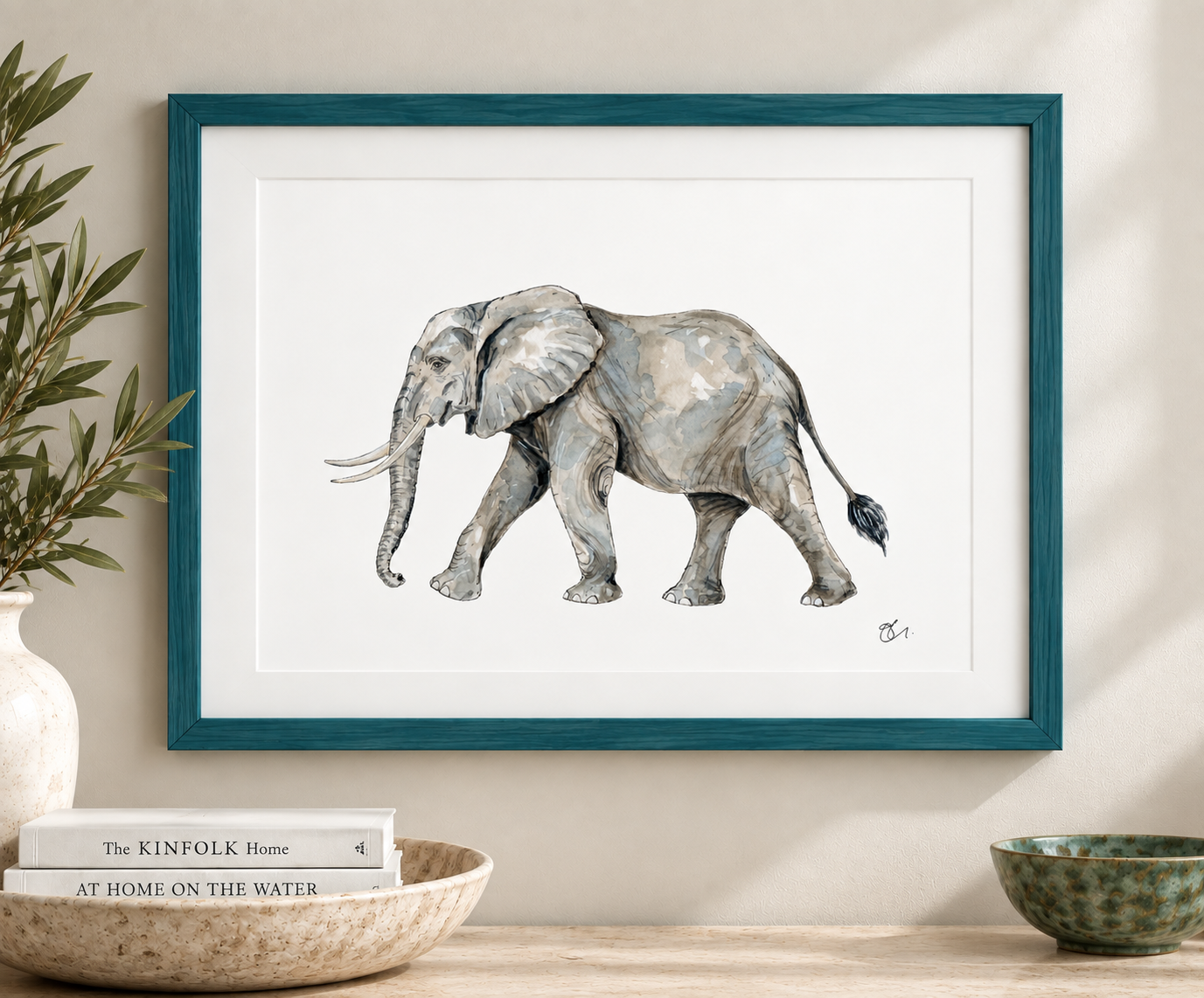 Watercolor painting of a gray elephant with large ears and tusks, framed in teal, hanging above a wooden table with potted plant, books, and bowls.