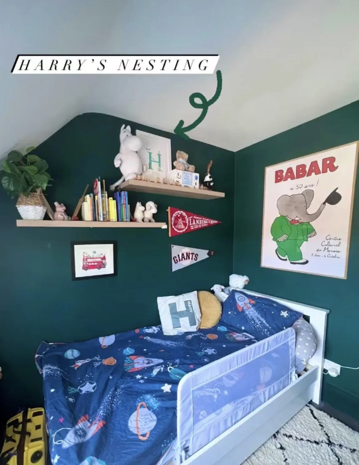 A children's bedroom with a space-themed bedspread, wall shelves with stuffed animals and books, and a green wall decorated with framed pictures, banners, and artwork including an elephant illustration.