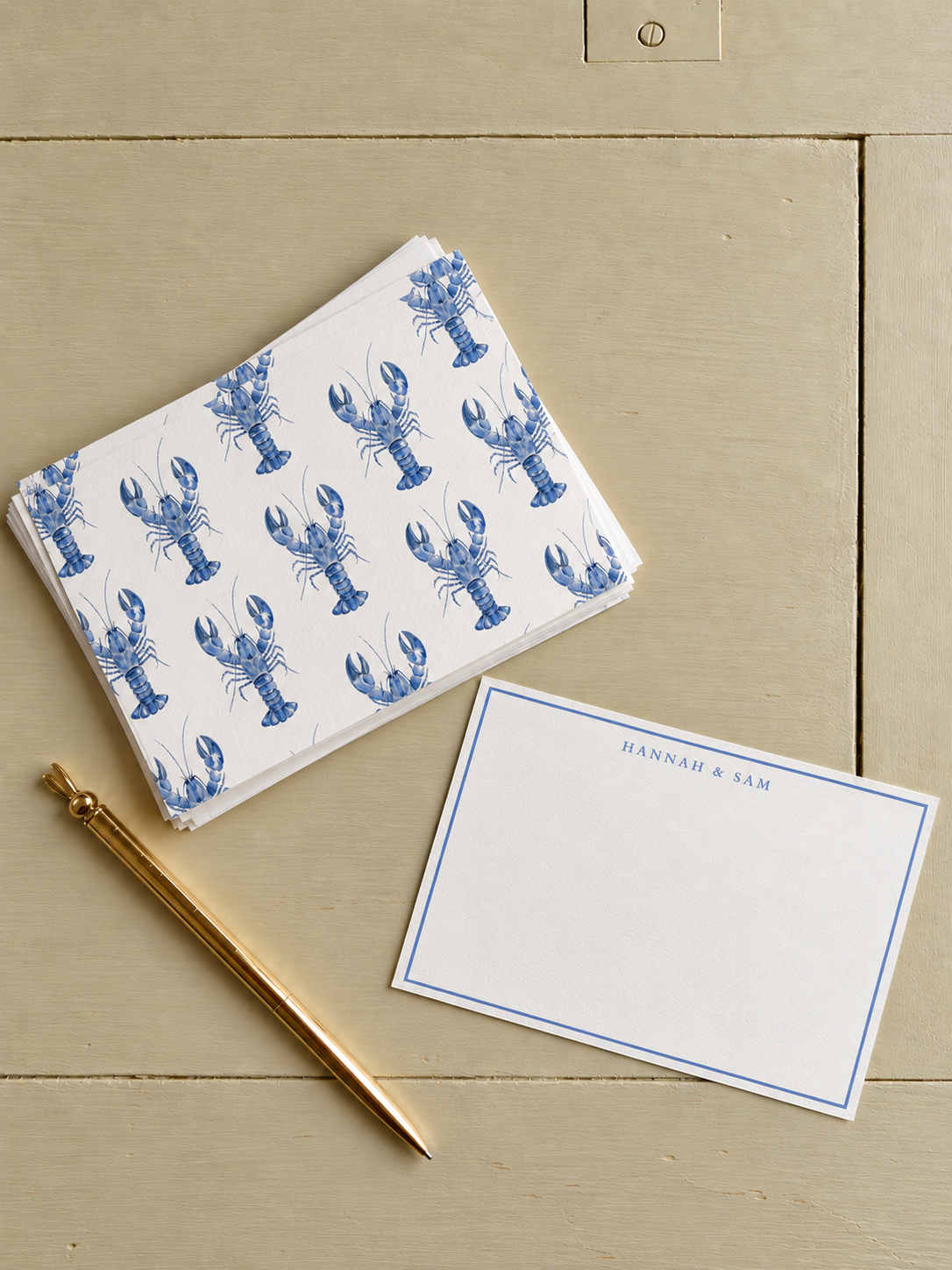 A stack of napkins with blue lobster prints, a blank notecard with 'Hannah & Sam' at the top, and a gold pen on a beige wooden surface.