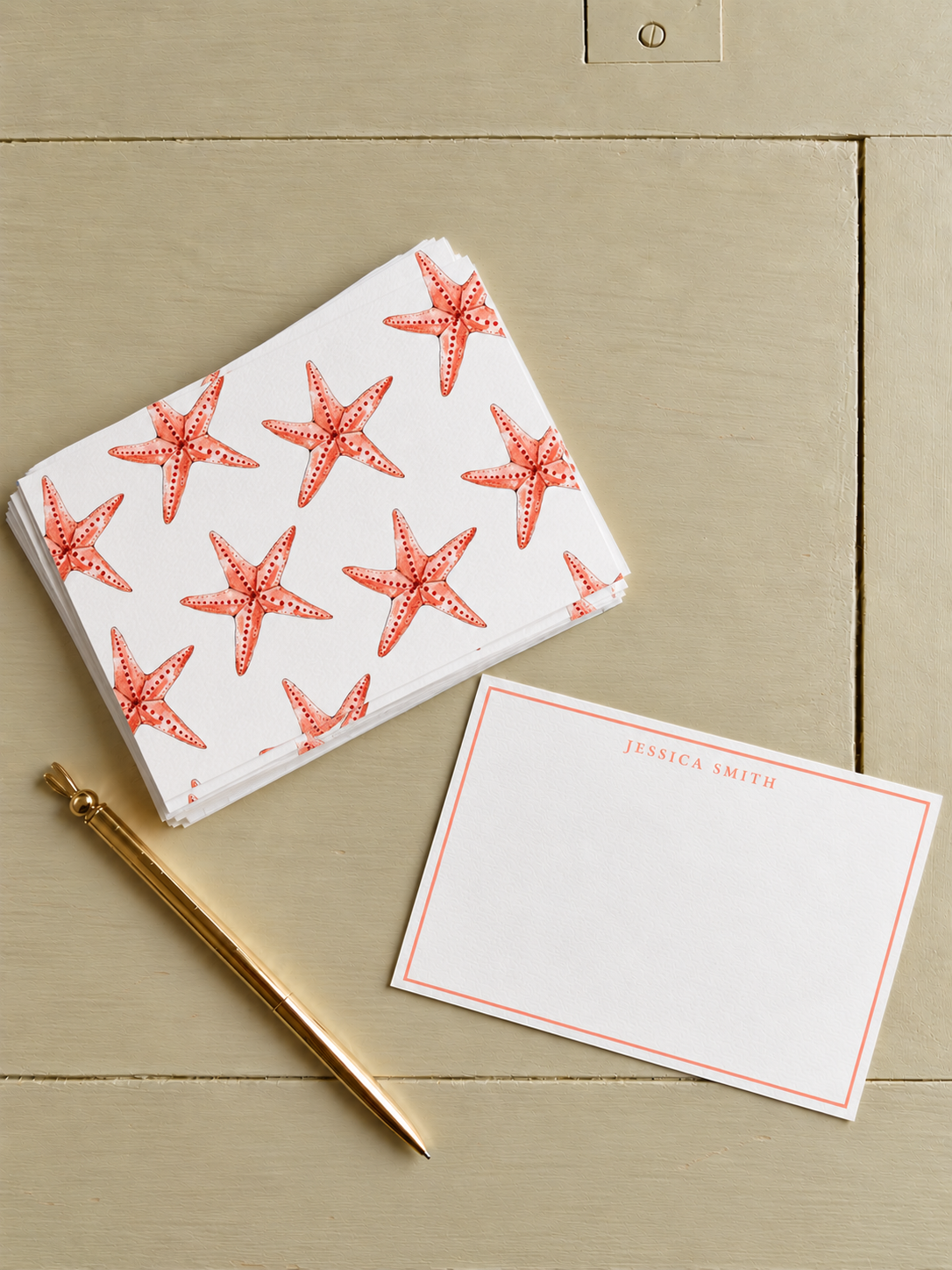 A stack of white napkins with starfish patterns, a blank note card with a pink border and "Jessica Smith" printed on it, and a gold pen, all on a beige wooden surface.