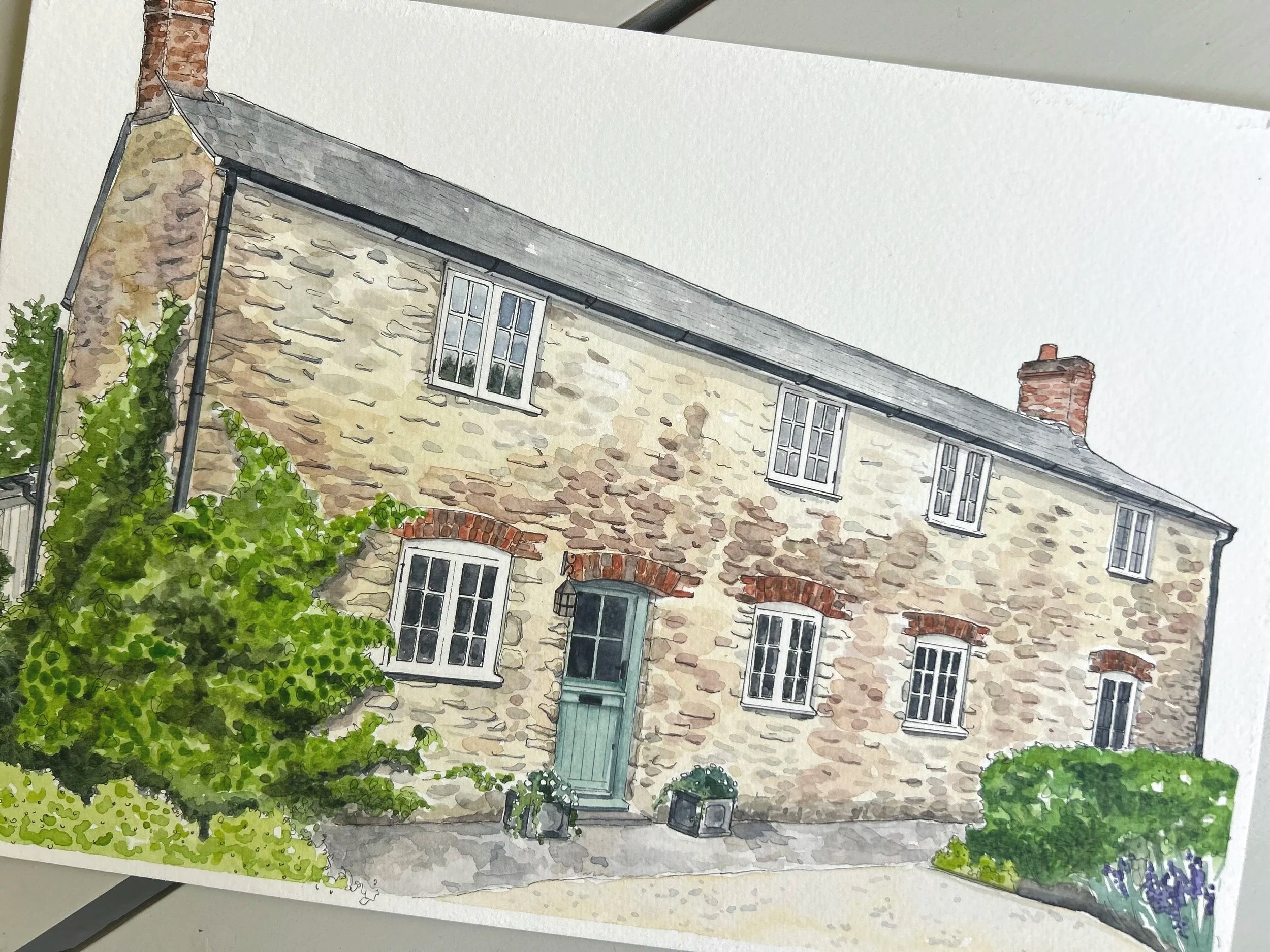 Watercolor painting of a stone cottage with a grey roof, large windows, and a brick chimney, surrounded by green bushes and trees.