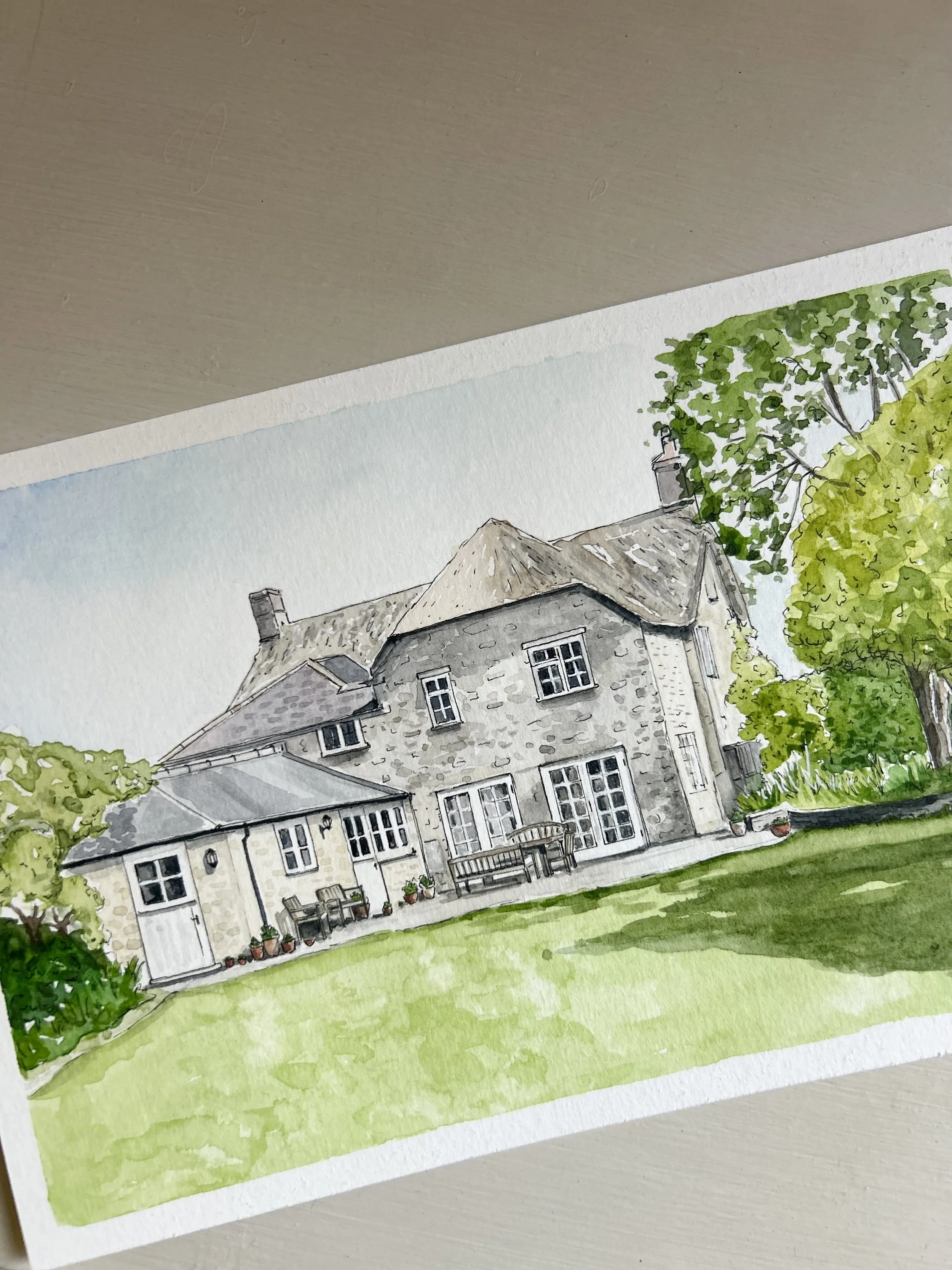 Watercolor painting of a large stone house with a slate roof, multiple chimneys, and a patio with chairs and potted plants, surrounded by greenery and trees.