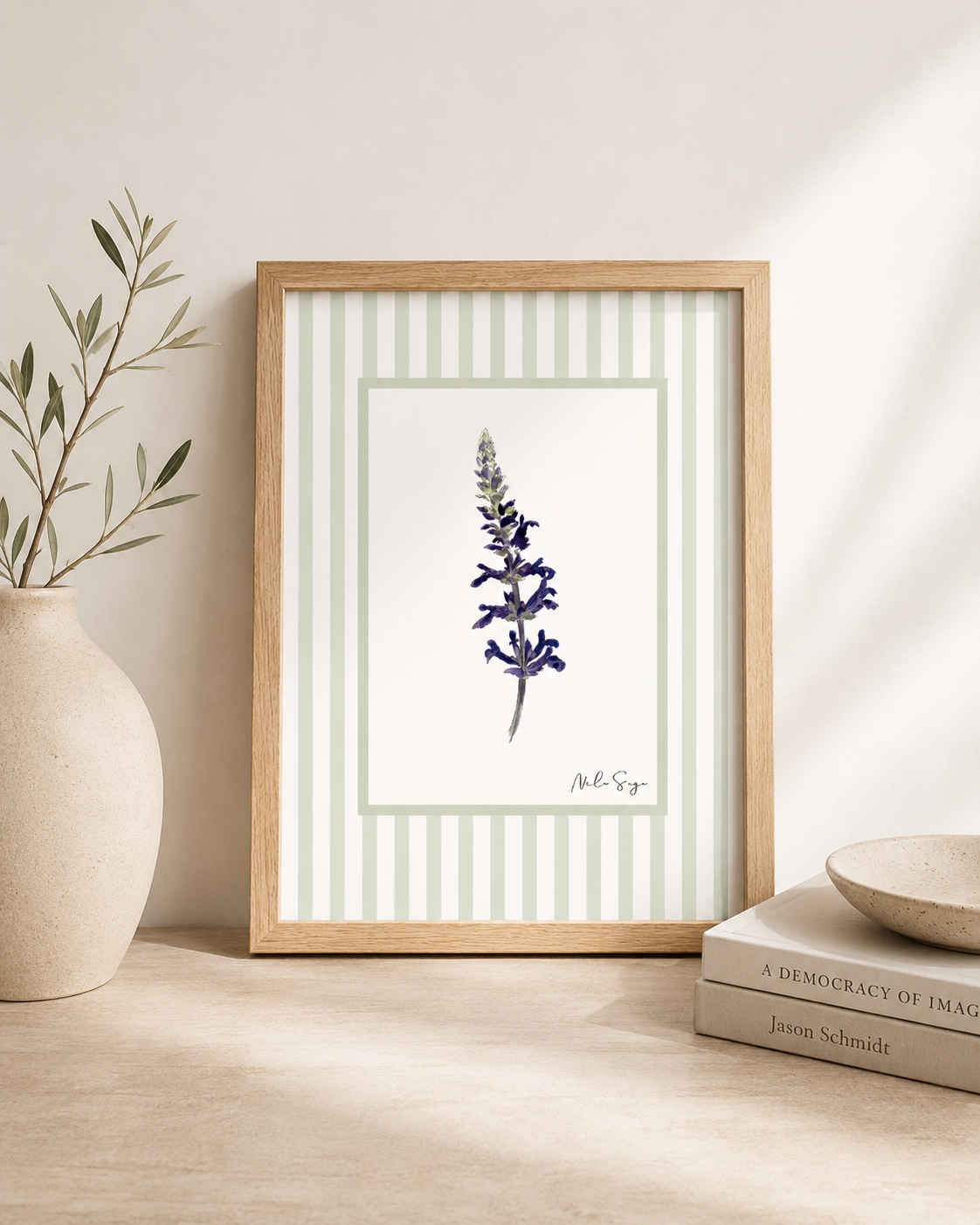 A framed botanical illustration of a purple flower, leaning against a white wall, with a potted plant on the left and books on the right.