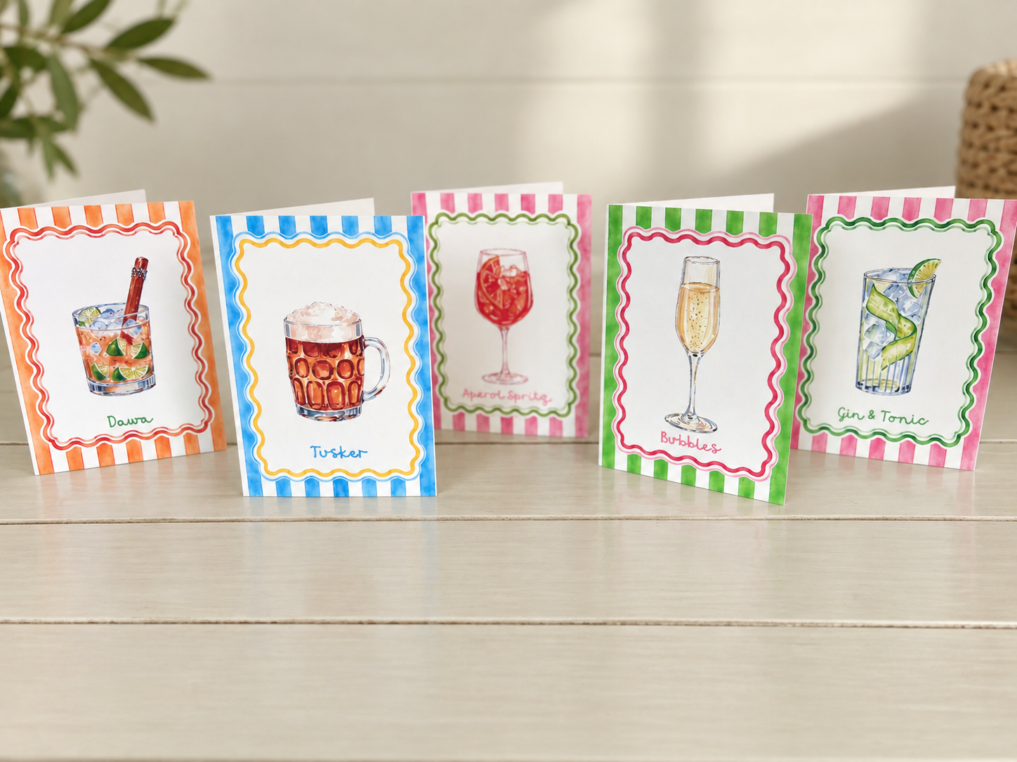 Colorful illustrated cocktail and drink recipe cards displayed on a light wooden surface.