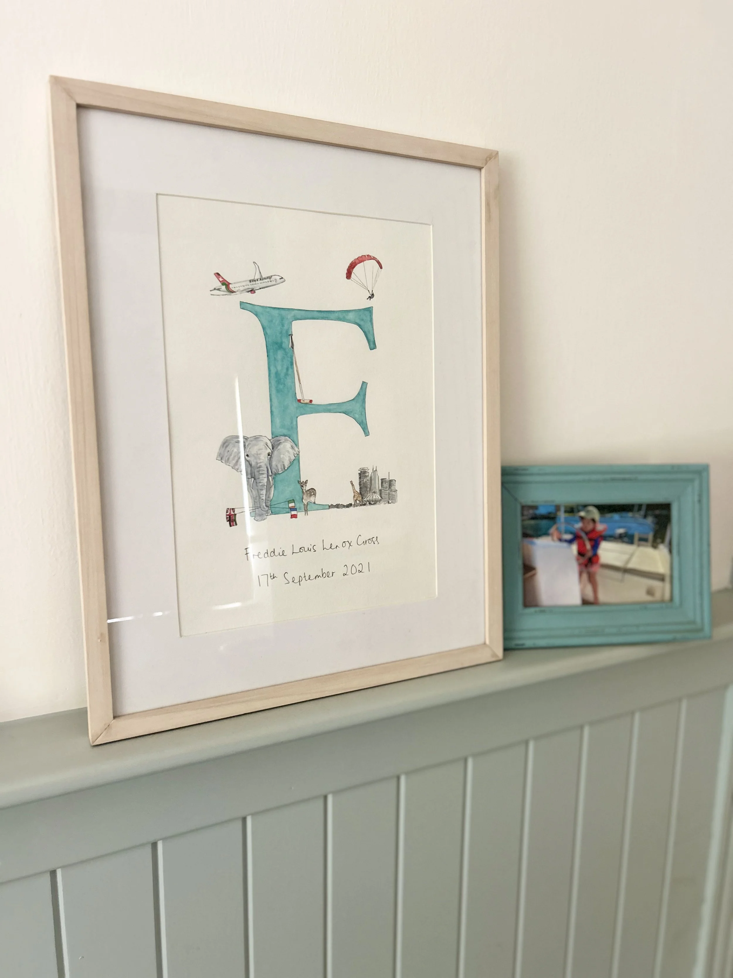A framed artwork featuring a large letter 'E' with various illustrations including an airplane, a parachute, an elephant, and other small figures and objects, with handwritten text below reading "Freddie Louis Hen Ox Cross 17th September 2021." A smaller photo of a child on a boat is partially visible in a blue frame in the background.