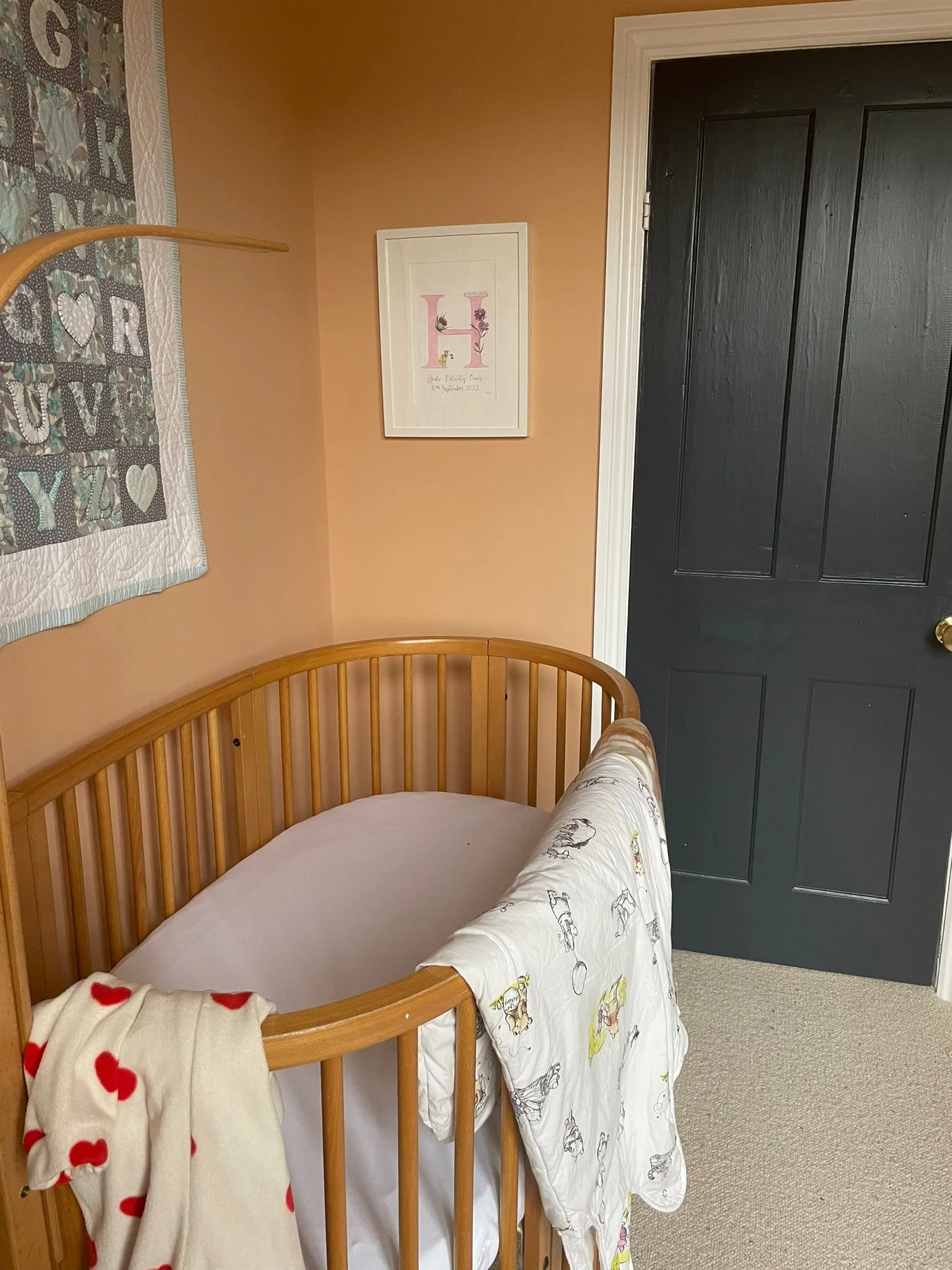 A view of a baby's crib with a white sheet and a blanket with red hearts, situated in a corner of a room next to a black door. A framed artwork with the letter 'H' decorated with flowers hangs on the beige wall. Part of a quilt with the word 'LOVE' and hearts is visible on the left side.
