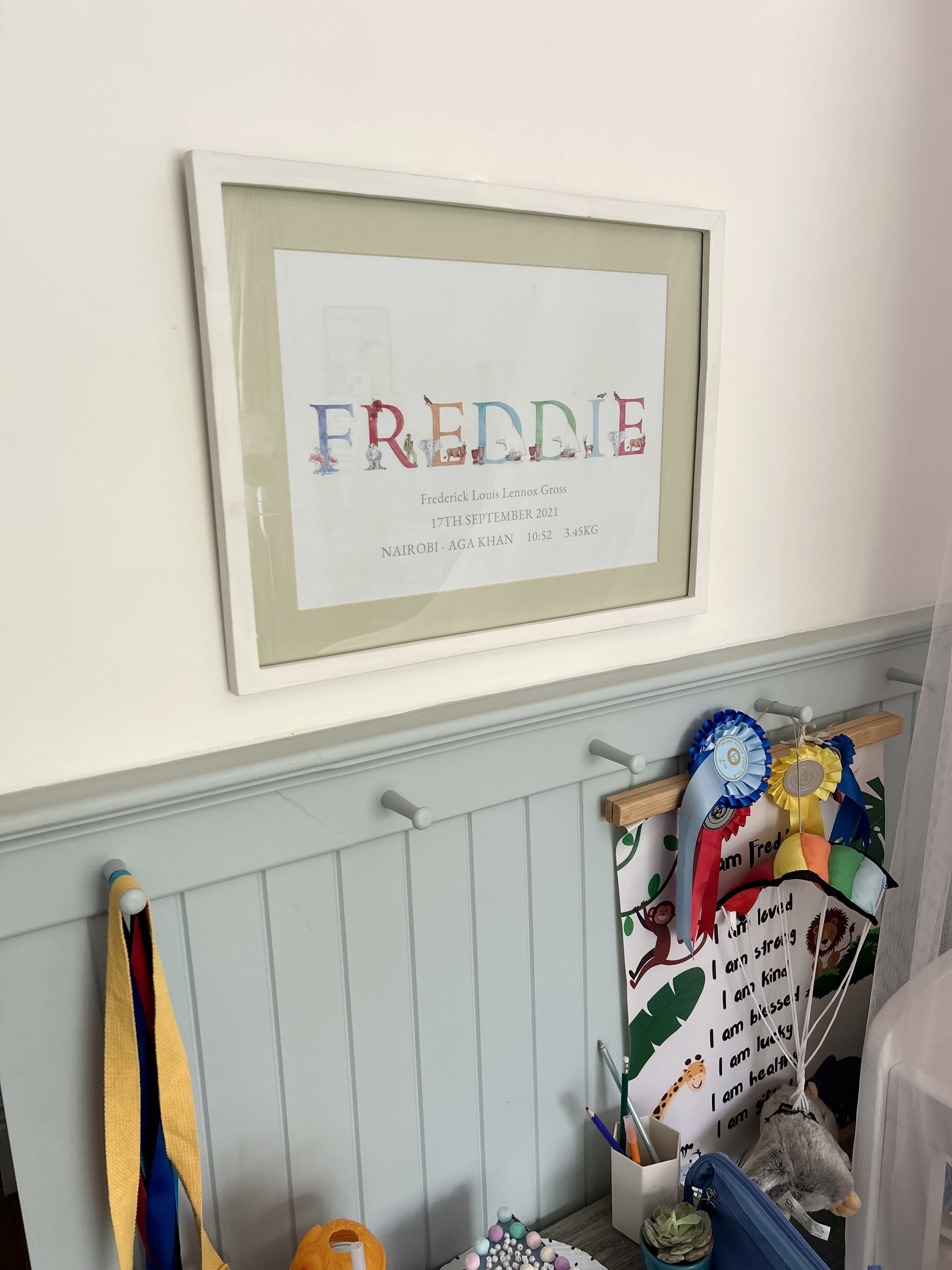 Framed birth announcement with the name Freddie, birth details, and colorful letters, hanging on a white wall above a white beadboard panel decorated with ribbons, toys, and various items.