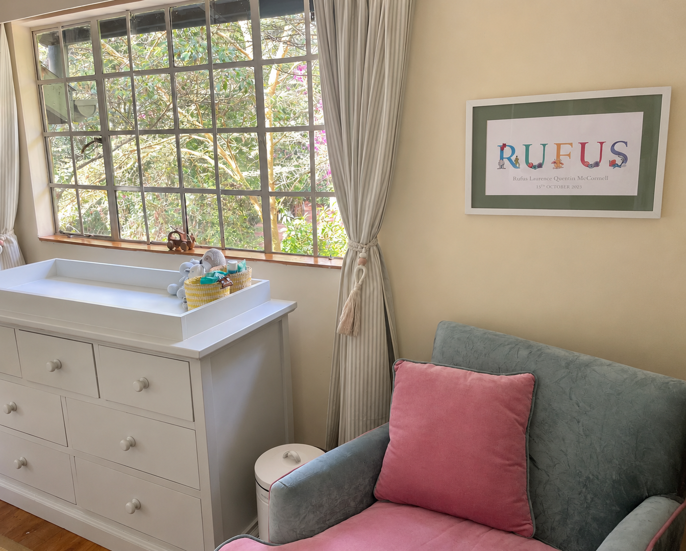 A cozy nursery room with a large window showing green tree leaves outside. There is a white dresser with a changing pad on top, decorated with stuffed animals and baby care items. A pink cushion is on a soft green armchair with a pink pillow. A framed colorful artwork saying 'RUFLUS' is on the wall.