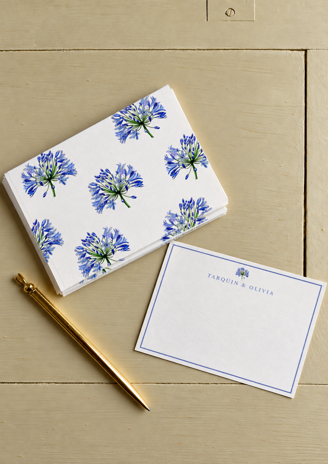 A set of note cards with a blue floral design, a white envelope with blue text and floral detail, and a gold pen on a beige wooden surface.