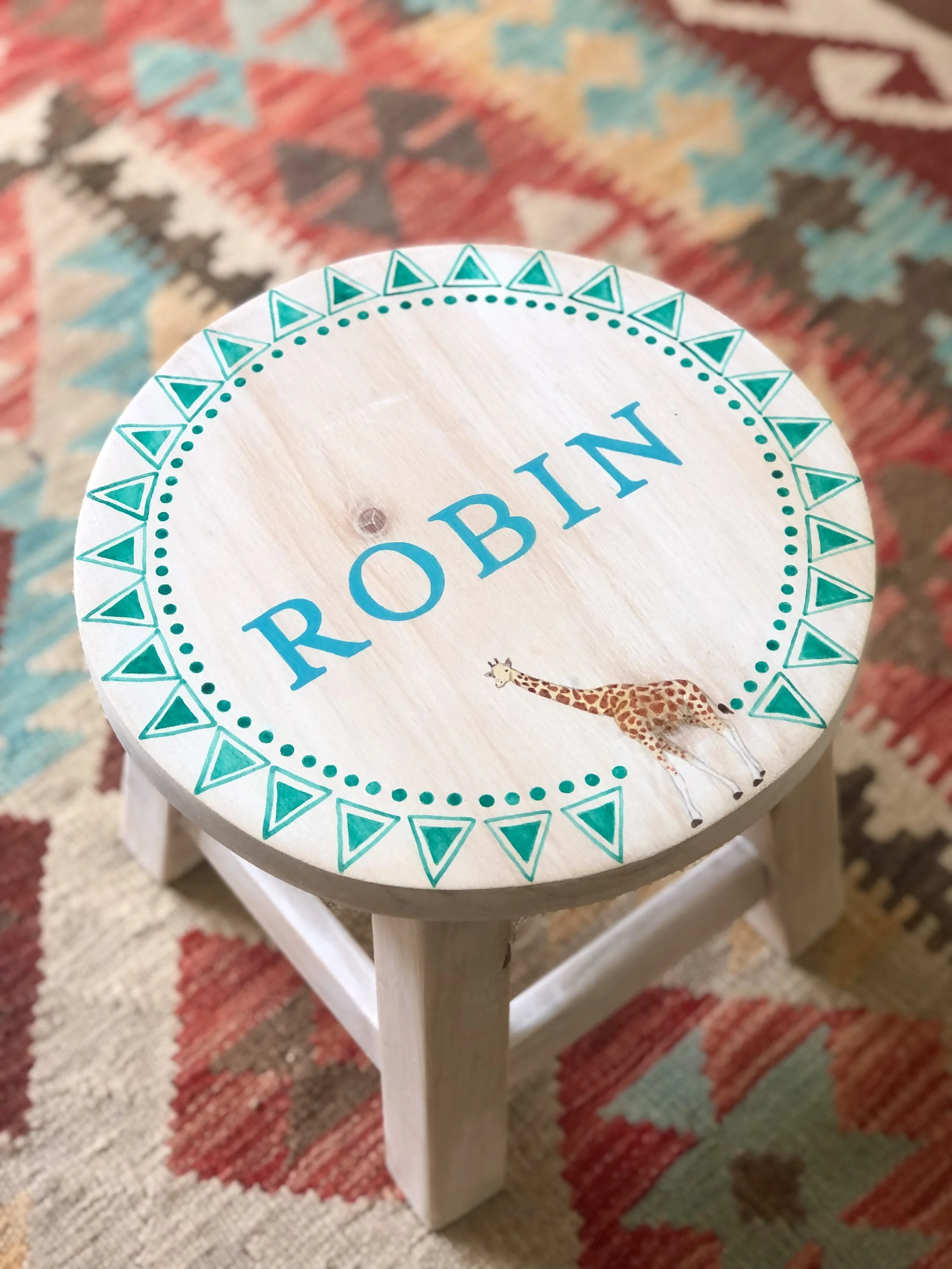 Round wooden stool with the word 'ROBIN' and a small giraffe illustration, decorated with a pattern of triangles around the edge, placed on a colorful patterned rug.