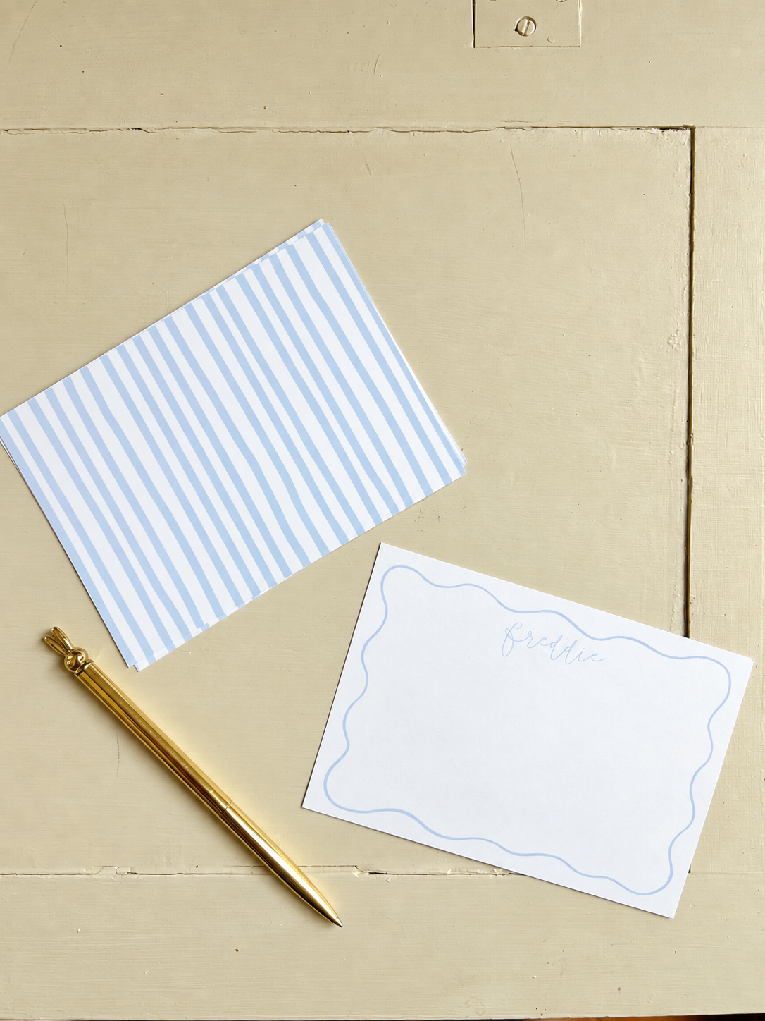 A gold pen, a blue and white striped envelope, and a white card with a blue border and the word 'freddie' written in cursive, arranged on a beige wooden surface.