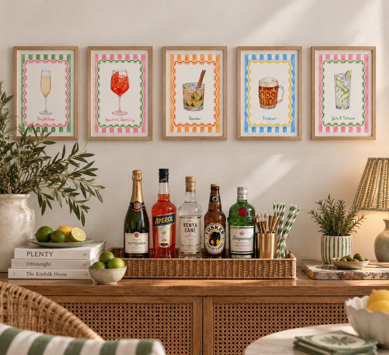 Decorative bar area with framed colorful cocktail illustrations on the wall, bottles of alcohol, citrus fruits, and bar accessories on a wooden sideboard.