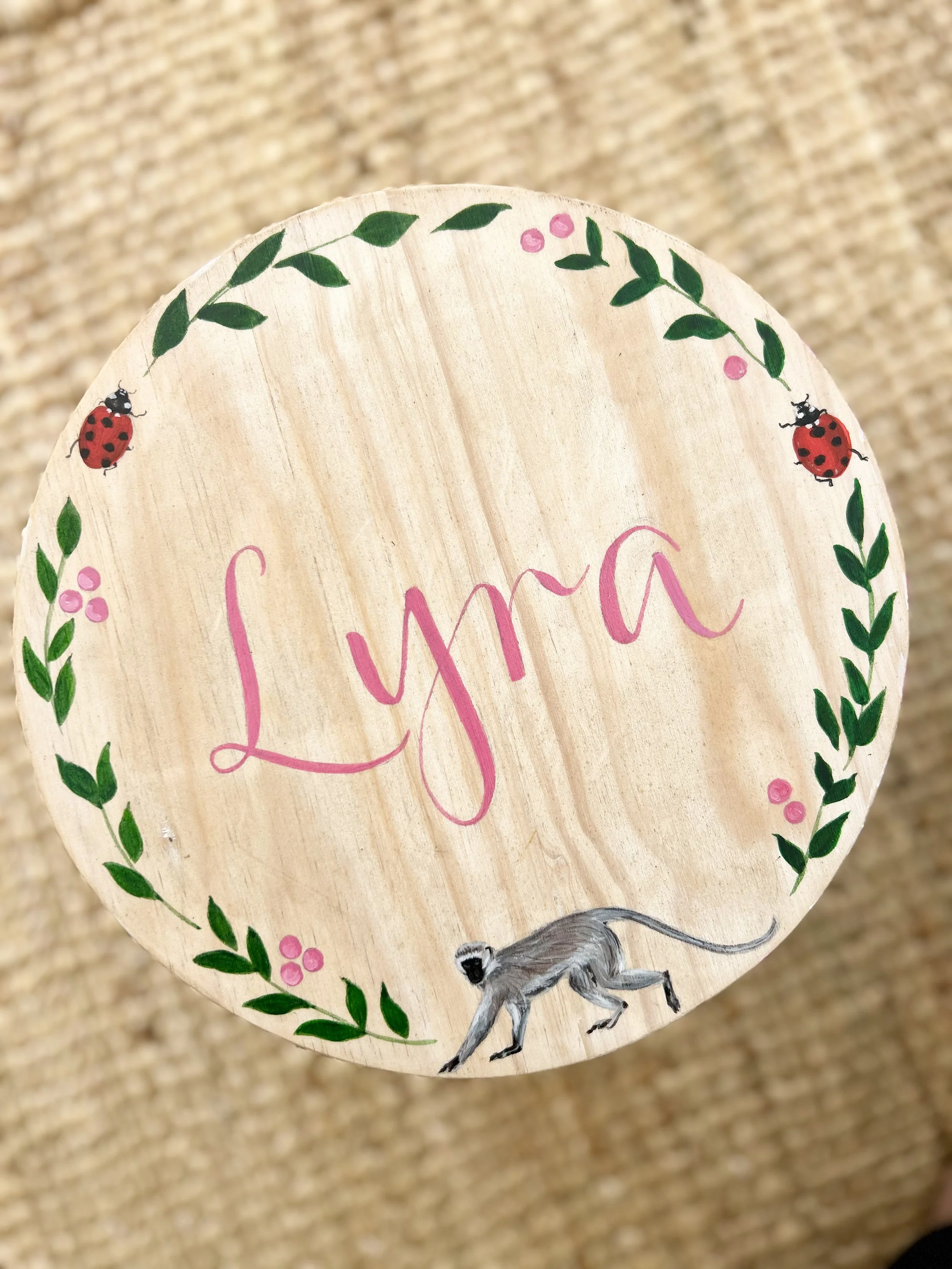 Close-up of a wooden sign decorated with green leaves, pink berries, and ladybugs, featuring the word 'Lysa' written in pink letters, with a small illustration of a running mouse at the bottom.