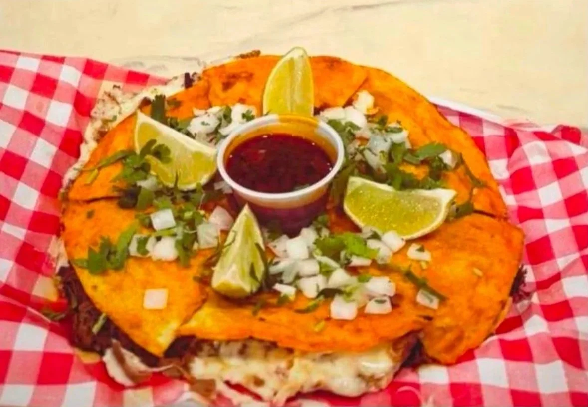 A plate of Mexican tostada with shredded meat, topped with chopped onions, cilantro, and lime wedges, served with a side of red salsa in a small cup on a red and white checkered paper.