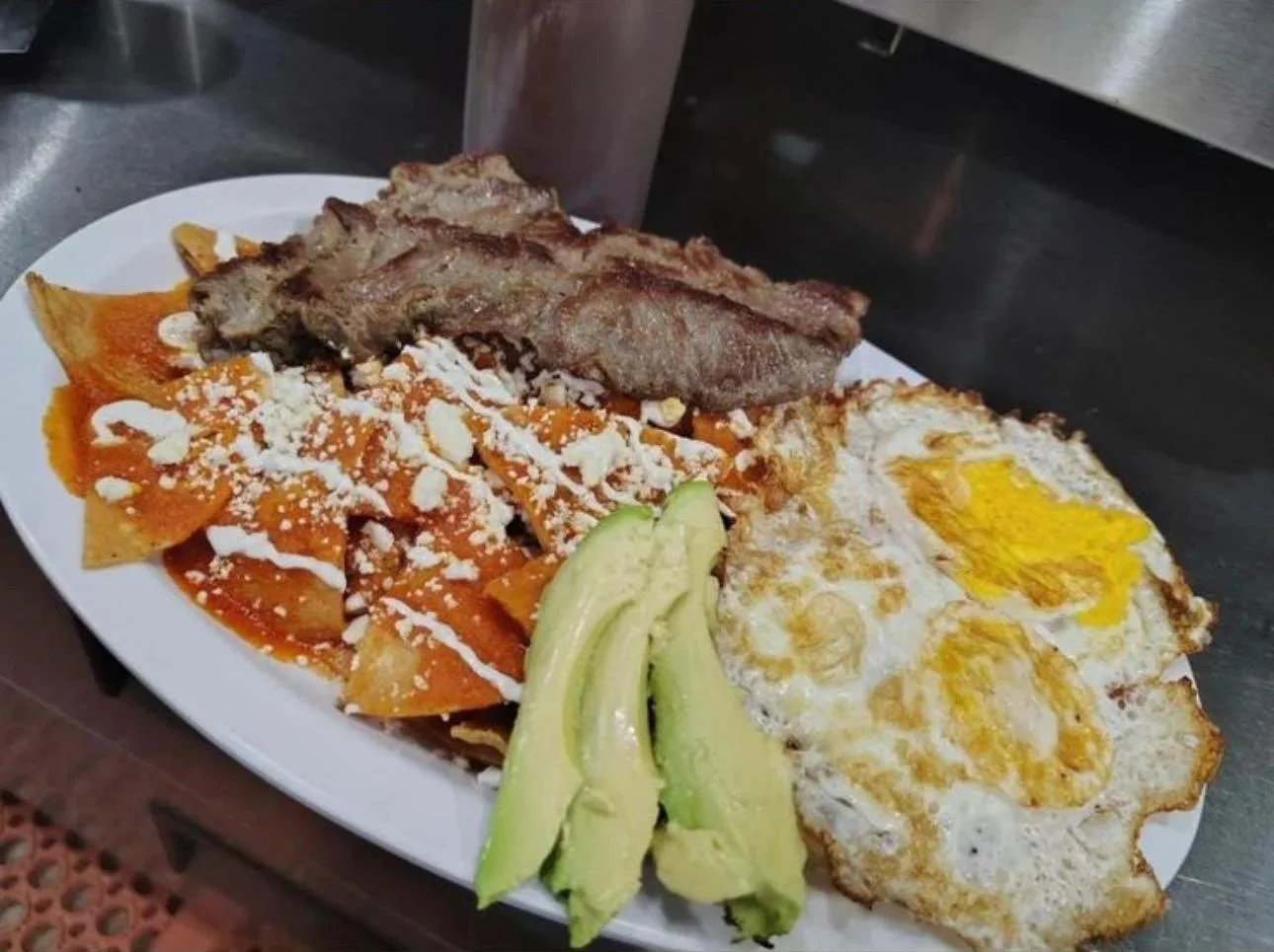 Plate of Mexican food including fried eggs, sliced avocado, roasted pork, and chilaquiles with cheese and red sauce.