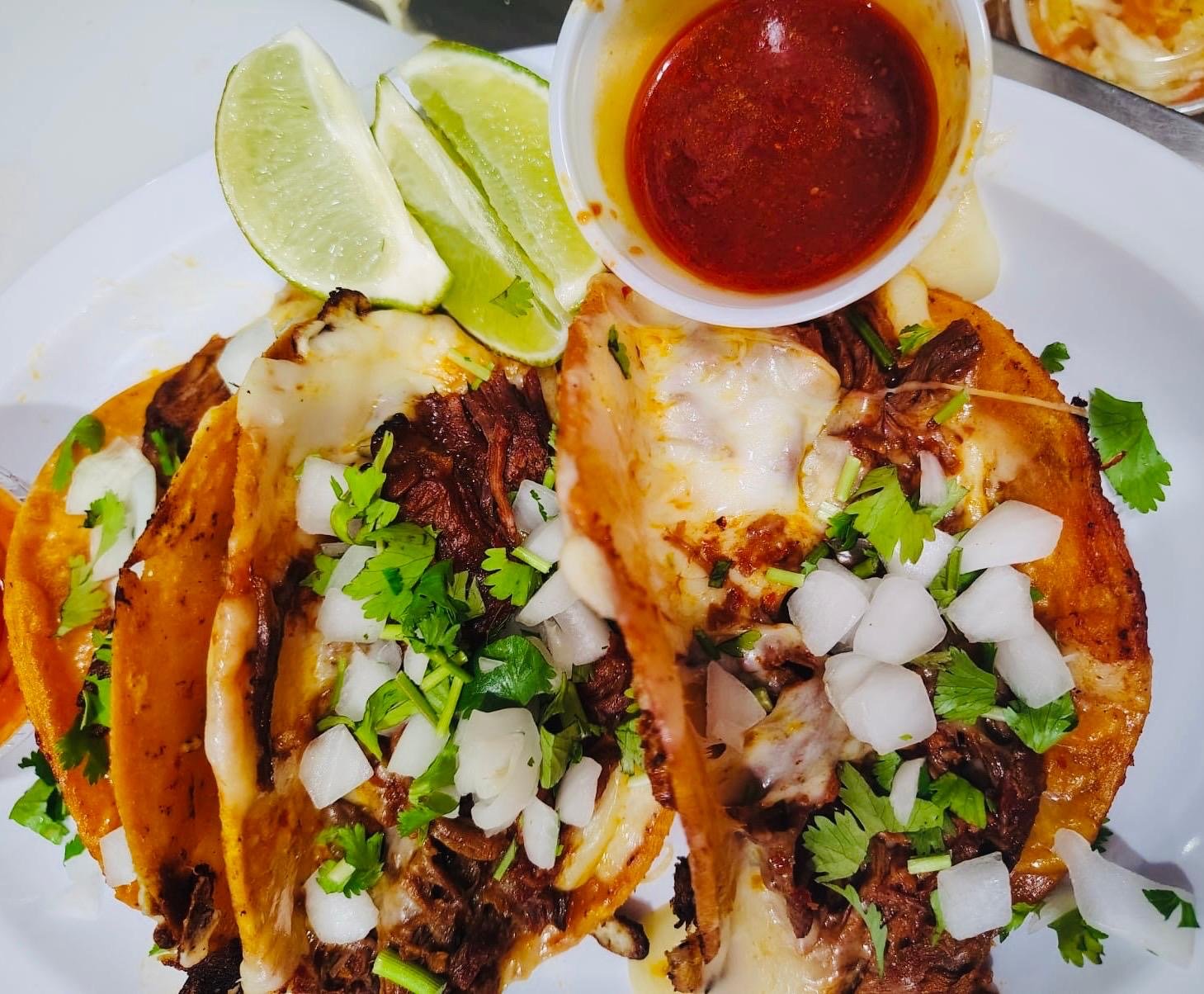 Close-up of three street-style tacos filled with shredded beef, onions, cilantro, and cheese, served with lime wedges and a small cup of red salsa on a white plate.