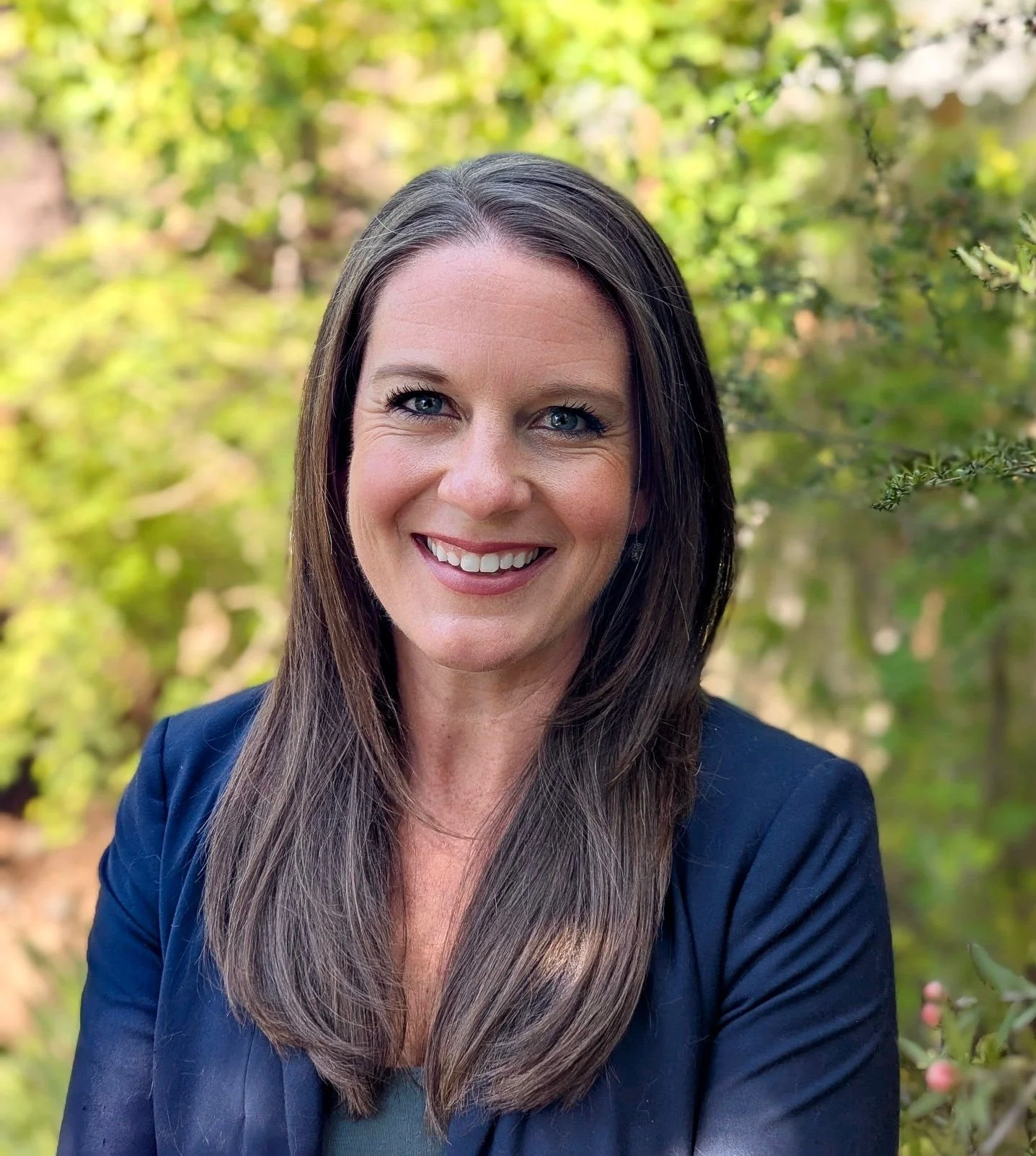 A woman with long brown hair, wearing a navy blazer, smiling outdoors with green foliage in the background.
