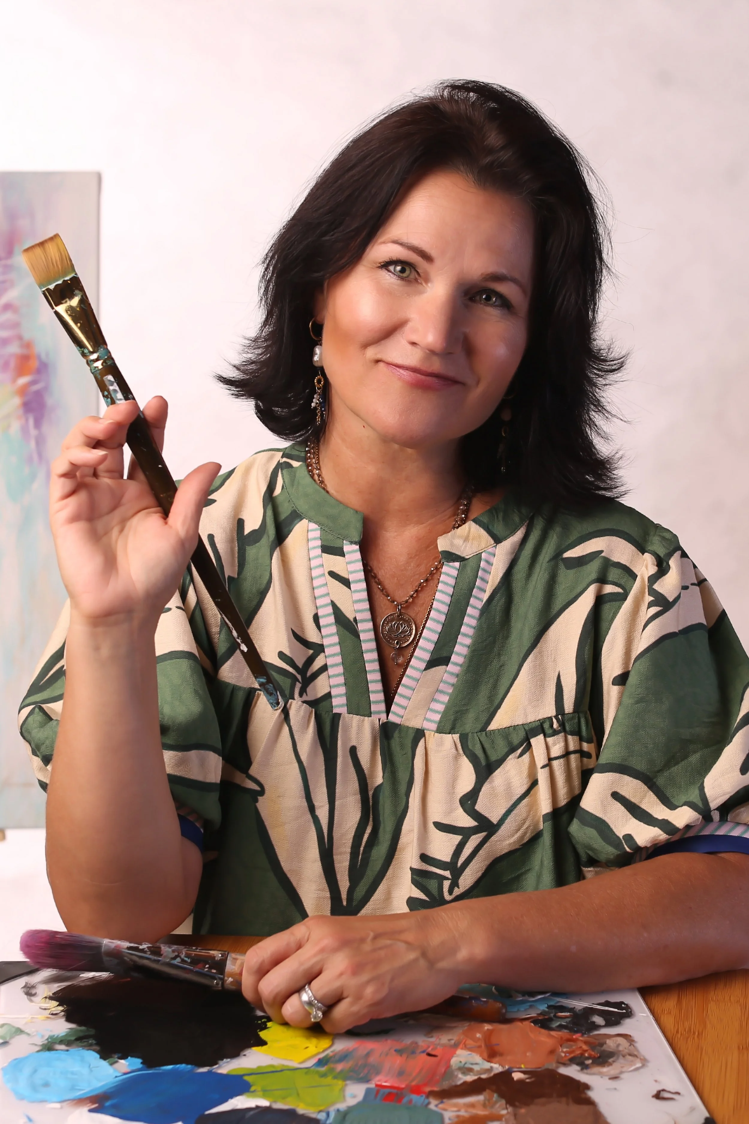 A woman with dark hair and earrings smiling while holding a paintbrush at a table with paint colors and a palette in front of her.