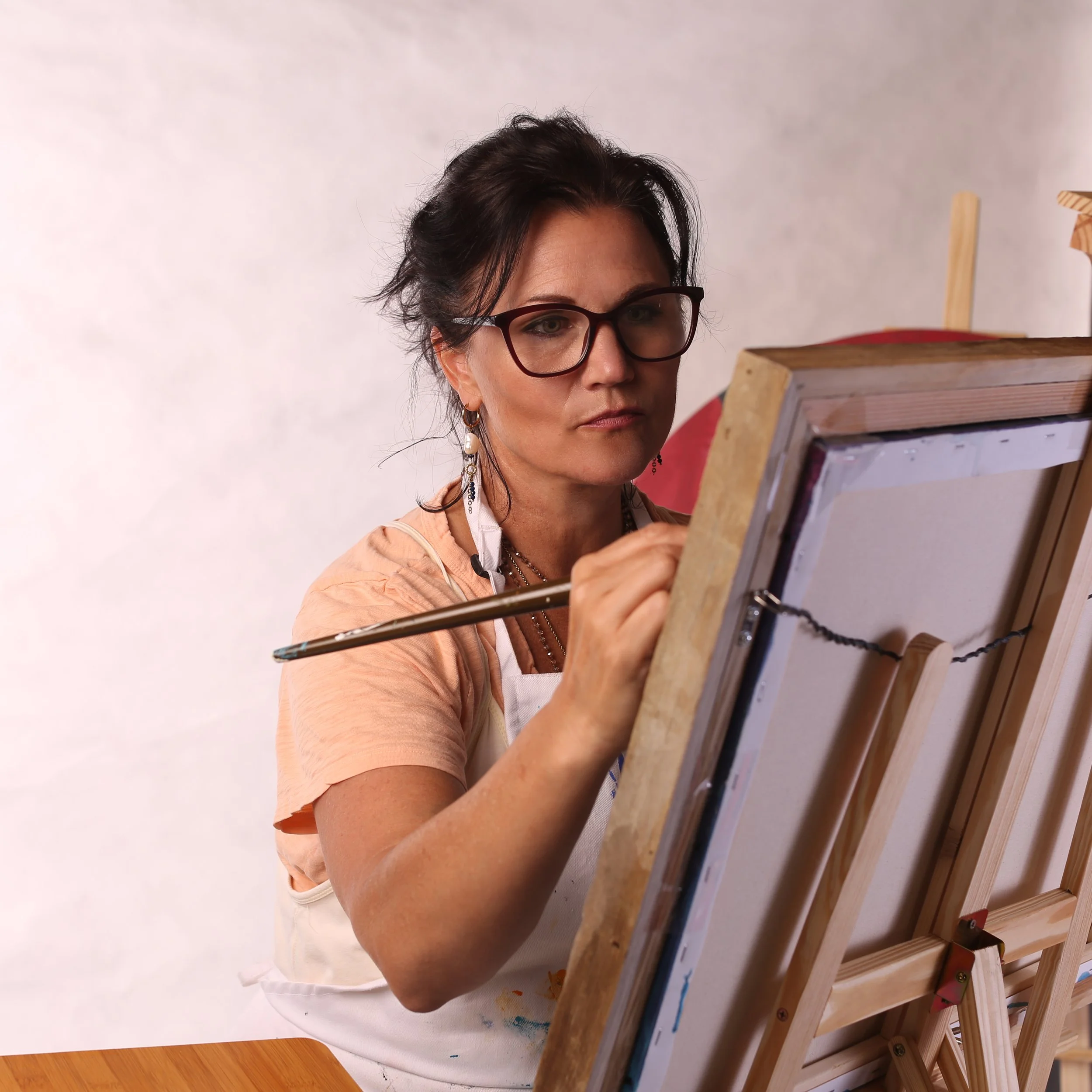 A woman with glasses and earrings looking at a canvass on an easel.