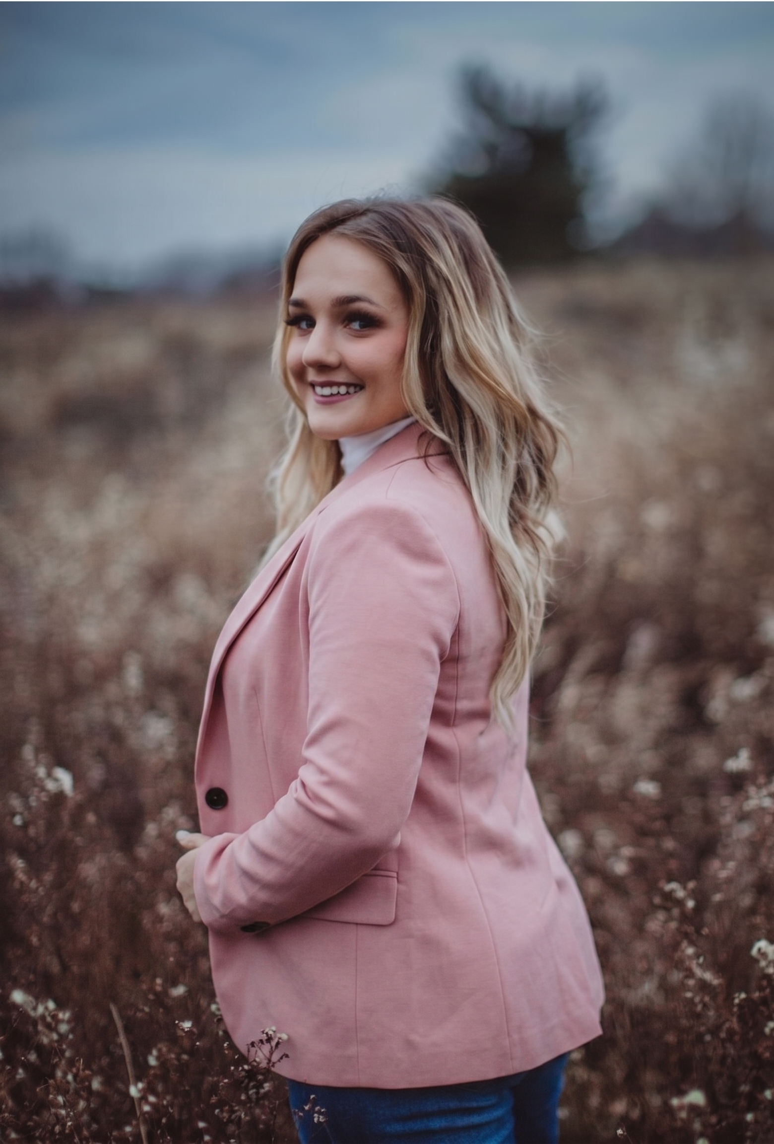 A young woman with long, wavy blonde hair wearing a pink blazer and blue jeans, smiling and looking at the camera outdoors in a field with dry plants and a cloudy sky in the background. Headshots for a local Peoria IL Realtor