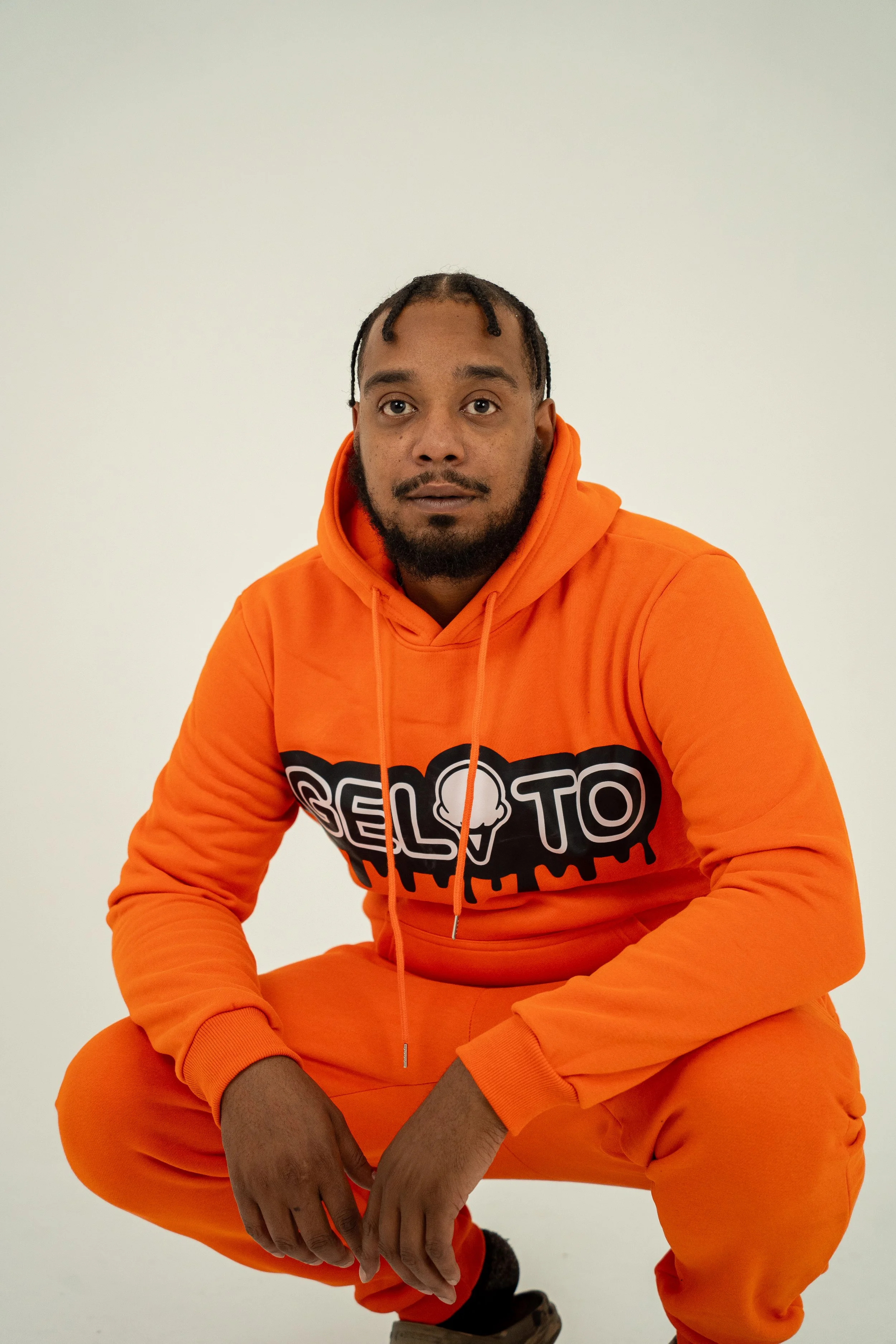 A man wearing a bright orange hoodie and matching orange pants, sitting in a squatting position with his hands clasped between his knees, against a plain light-colored background.