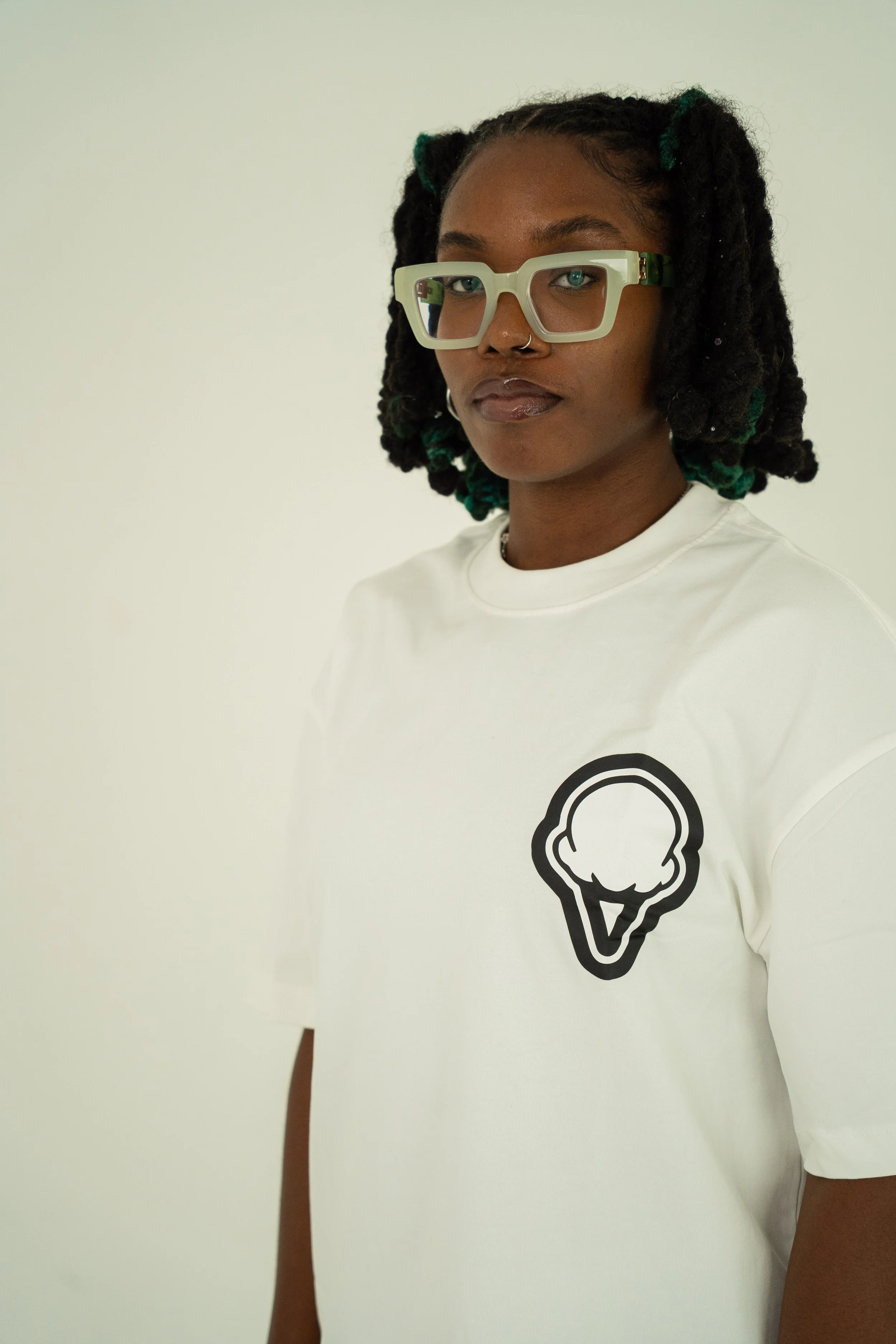 A young woman with glasses and dreadlocks wearing a white t-shirt with a black ice cream cone graphic, standing against a plain light-colored wall.