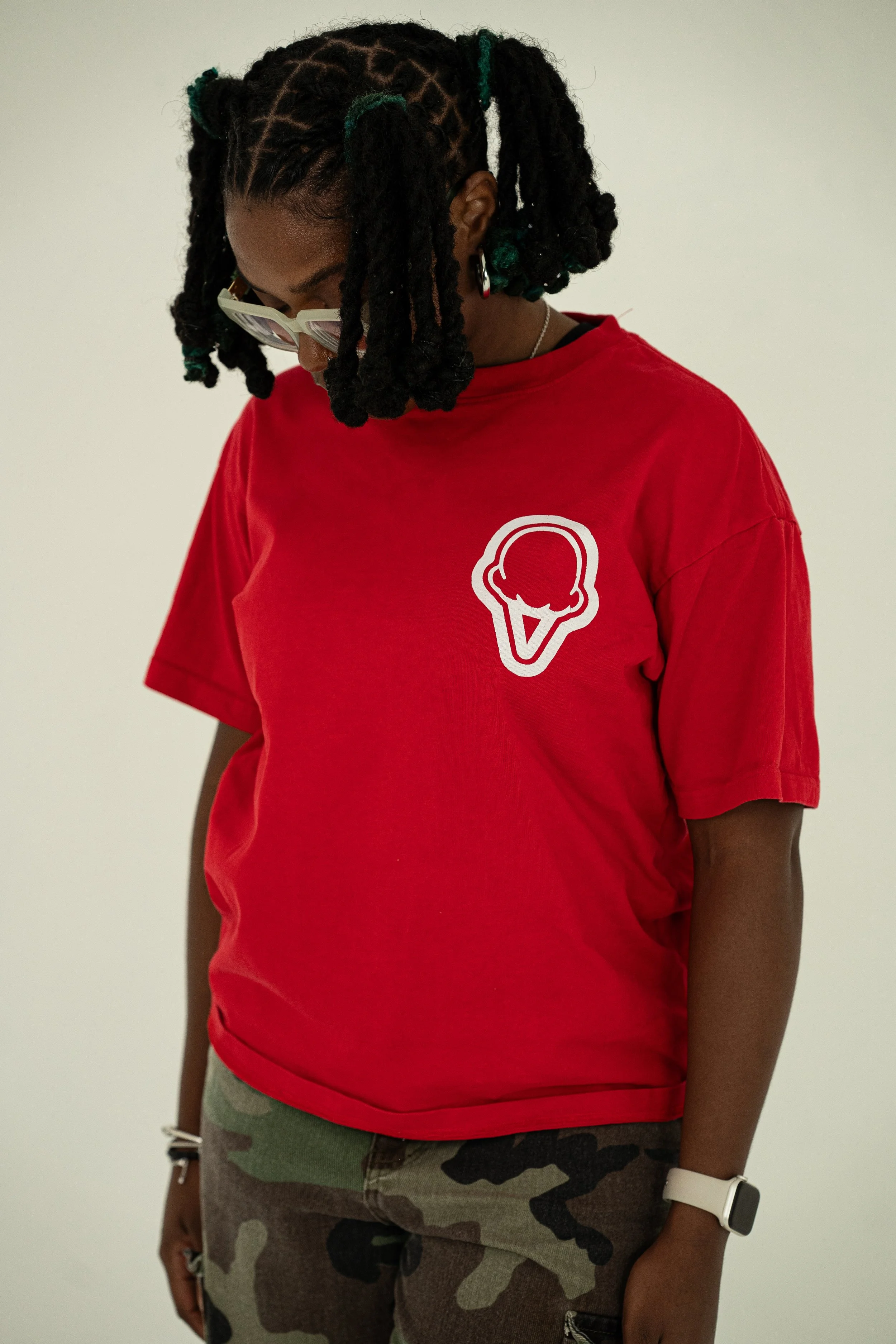 A person with dark skin and dreadlocks wearing glasses, a red t-shirt with an ice cream cone logo, camouflage pants, and a smartwatch, standing against a plain white background.