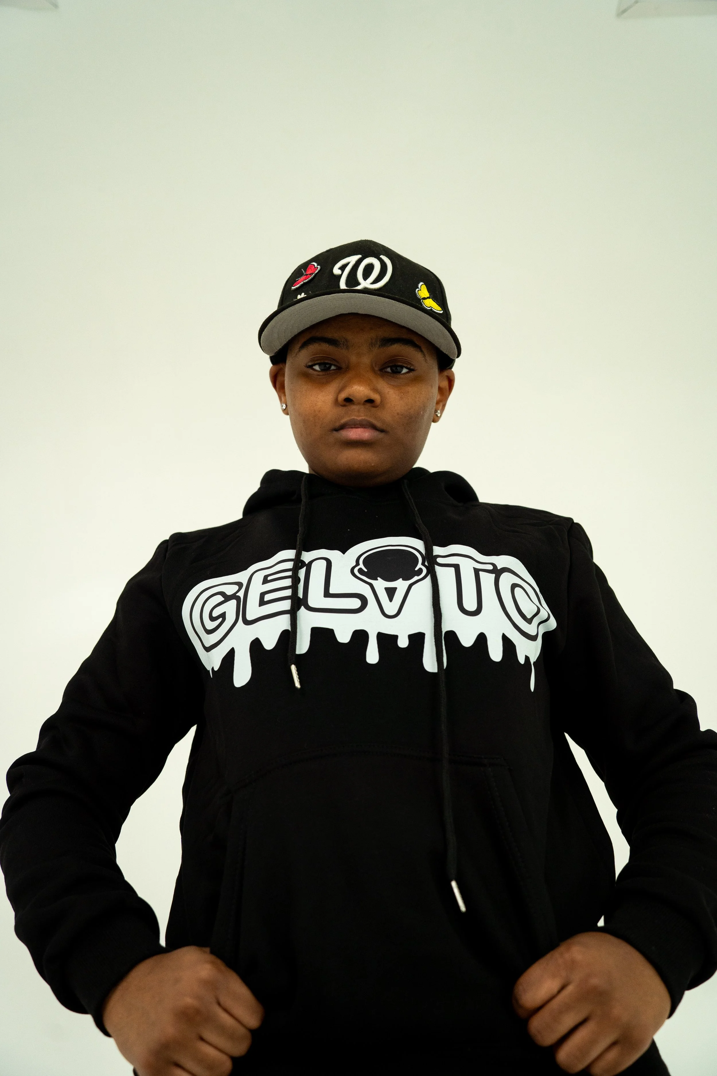 A young person with short hair wearing a black hoodie with a graphic and a black baseball cap with the Washington Nationals logo, standing against a plain white background.