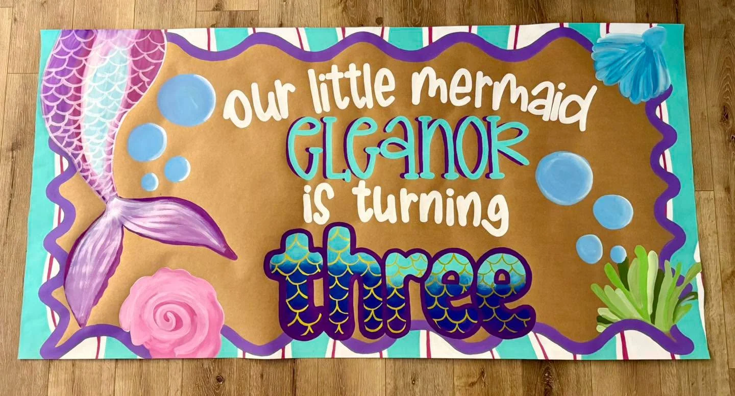 Decorated banner with mermaid theme featuring colorful text and illustrations of a mermaid tail, seashells, bubbles, and seaweed on a brown background.