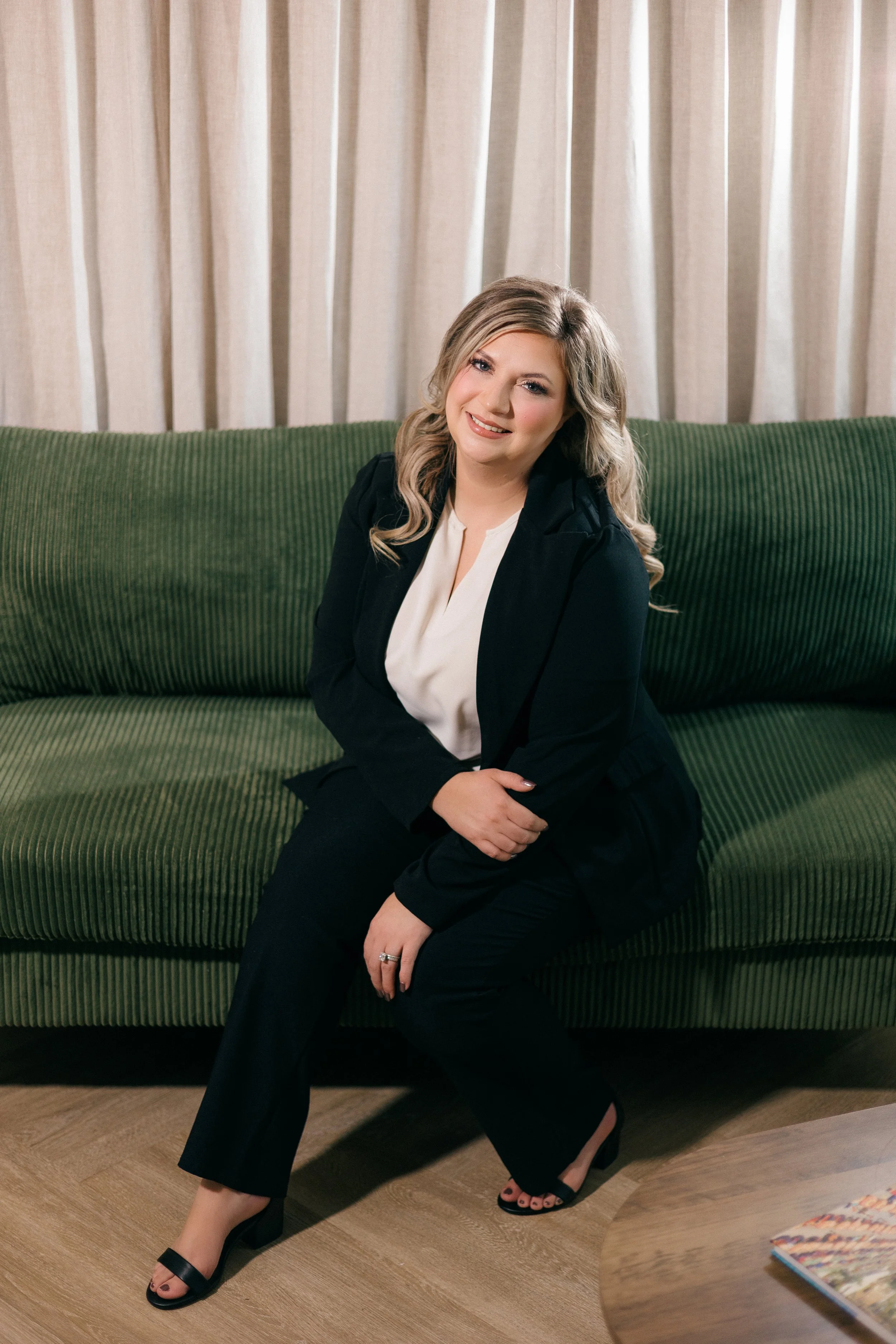 A woman sitting on a green sofa, dressed in a black blazer, white blouse, and black pants. She has blonde, wavy hair and is smiling at the camera. There is a curtain in the background and a magazine on a wooden table in the foreground.