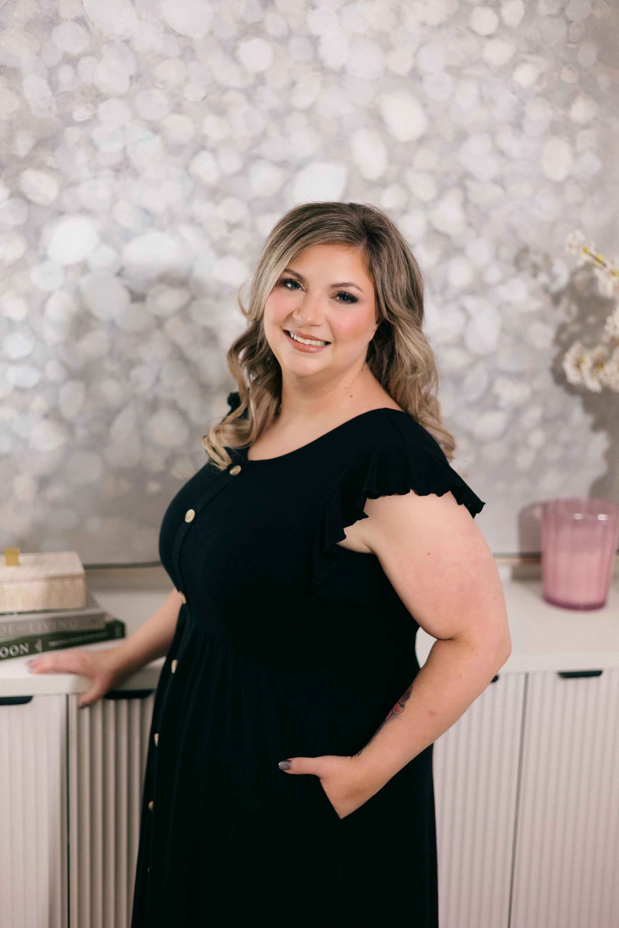 A woman with wavy blonde hair, makeup, and a black dress with ruffled sleeves and gold buttons, smiling while standing in a room with a white beadboard cabinet, a pink vase, and a textured gray and white background.