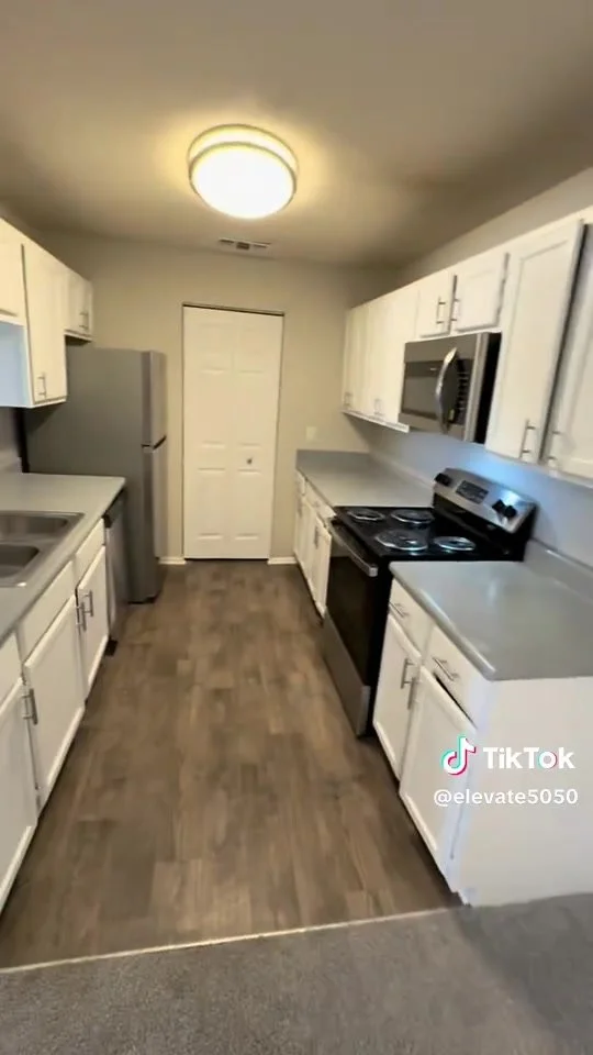 A narrow kitchen with white cabinets, gray countertops, a stainless steel refrigerator, stove, microwave, and wood-look flooring.