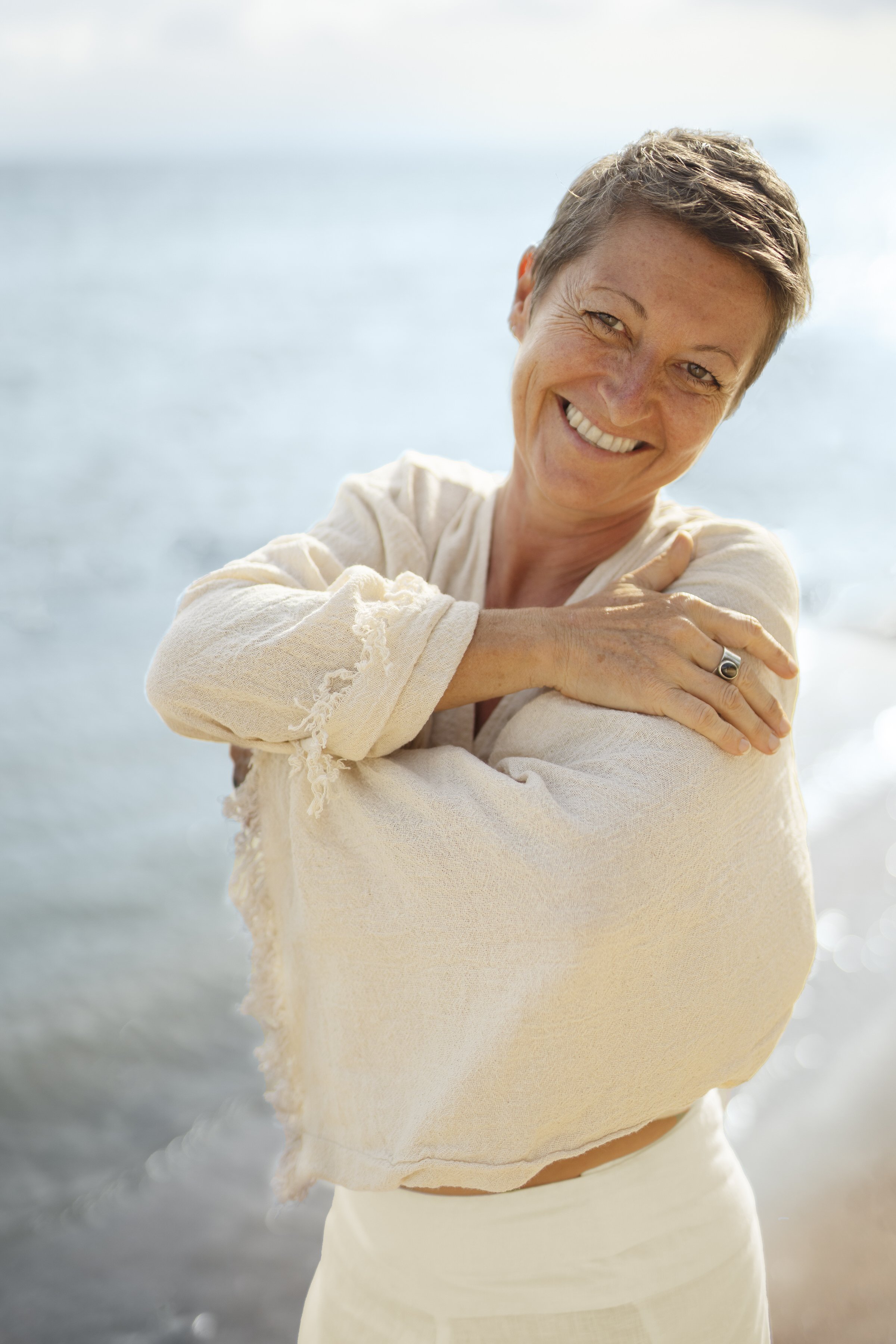 A woman with short brown hair, smiling, with a tan complexion, wearing a cream-colored shirt, standing on a beach with water in the background.