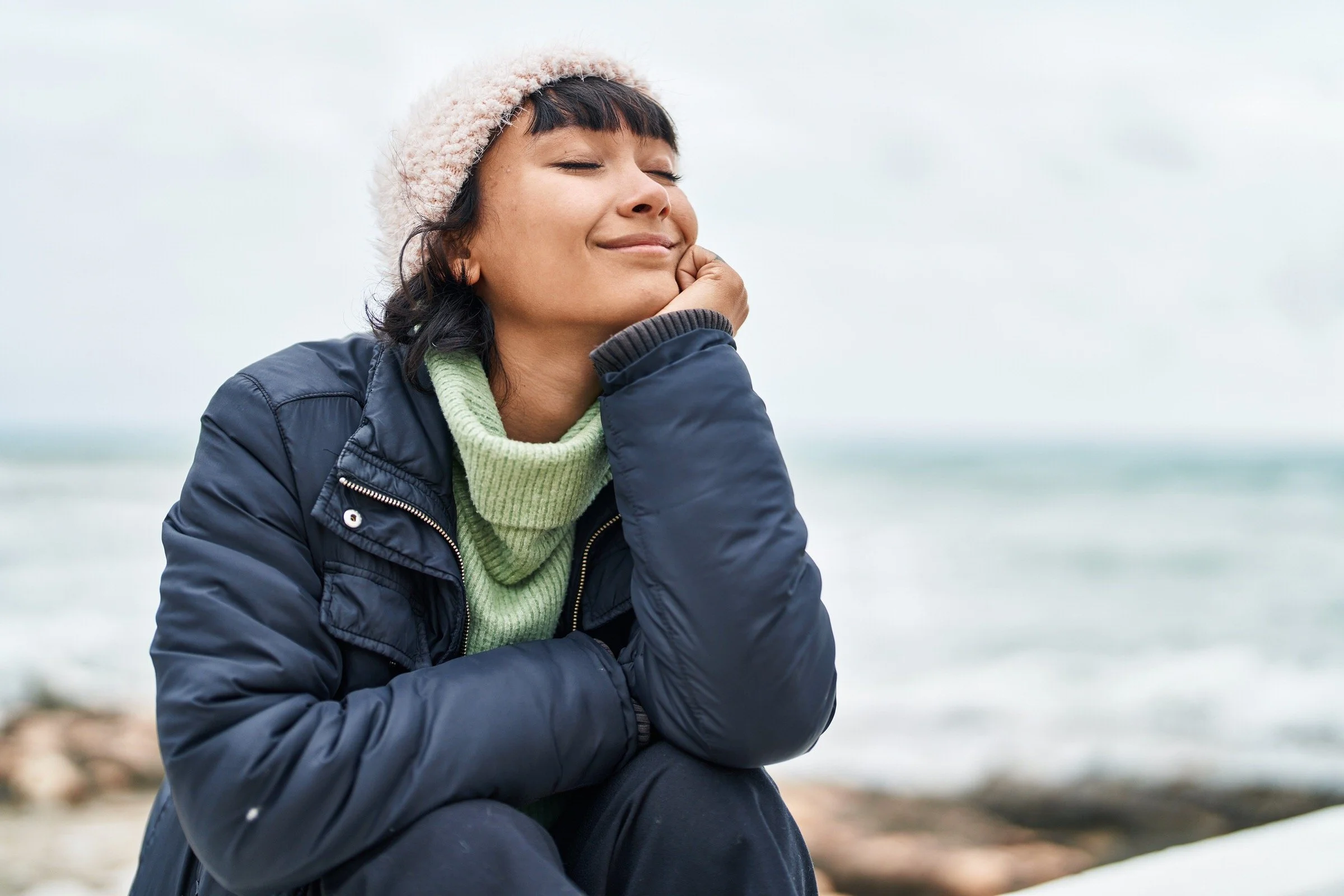 A woman with short dark hair, wearing a beige knit hat and navy jacket, standing outdoors with her eyes closed, smiling, and resting her chin on her hand against a blurred background of water and beach.