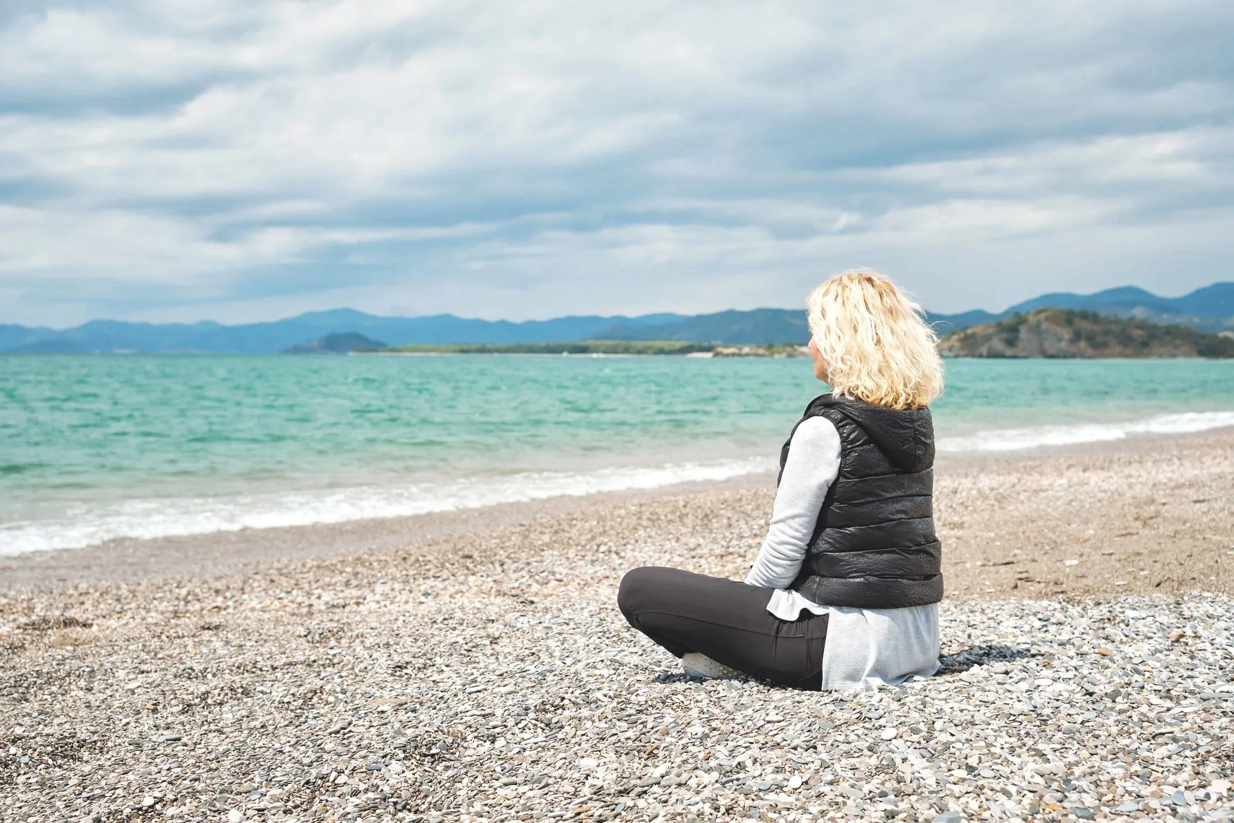 A woman with blonde curly hair, wearing a black vest and gray long-sleeve shirt, sitting cross-legged on a pebbled beach facing the water, with mountains and cloudy sky in the background.