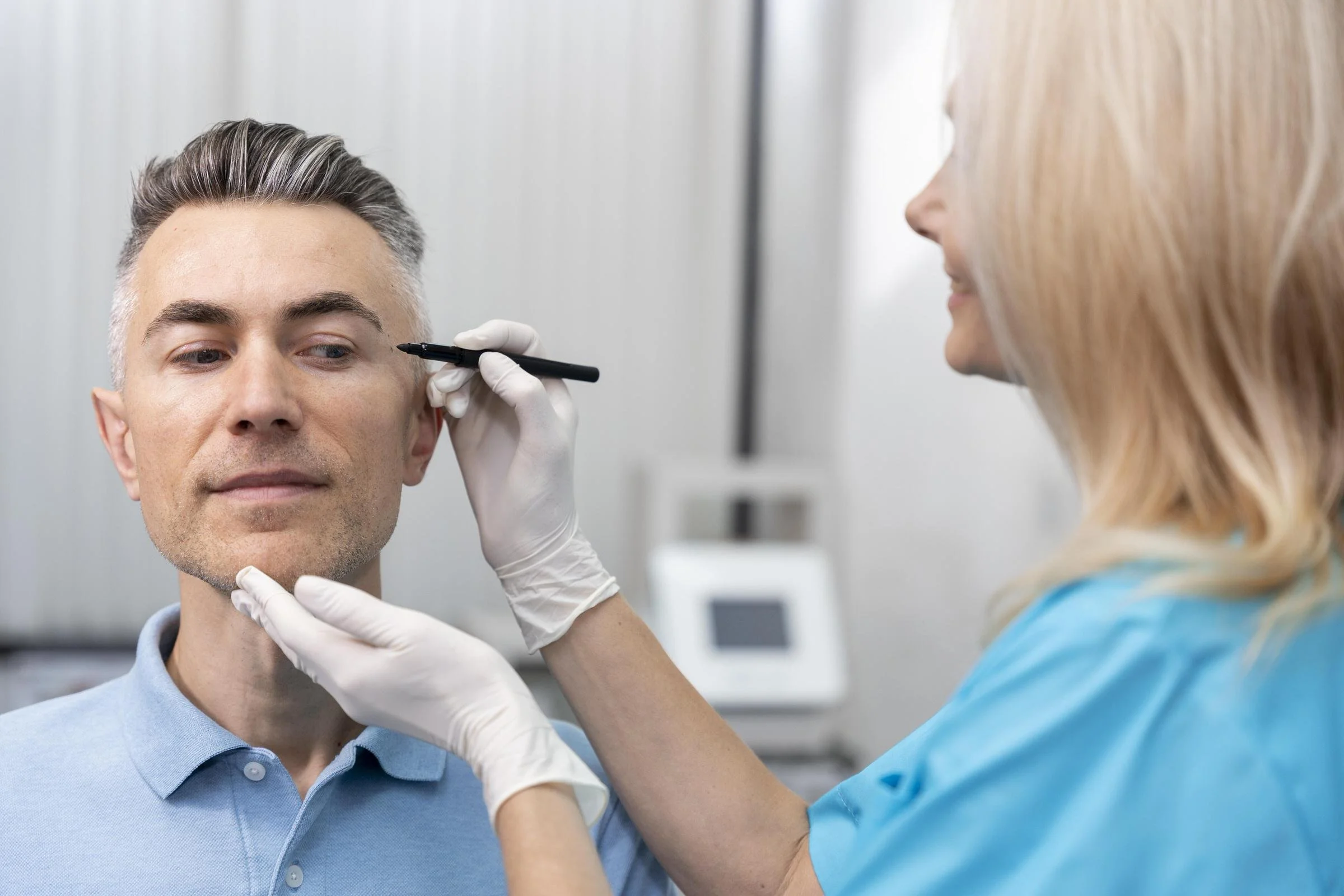 An osteopathic physician in blue scrubs and gloves drawing on a man's face during a cosmetic procedure in a clinical setting.
