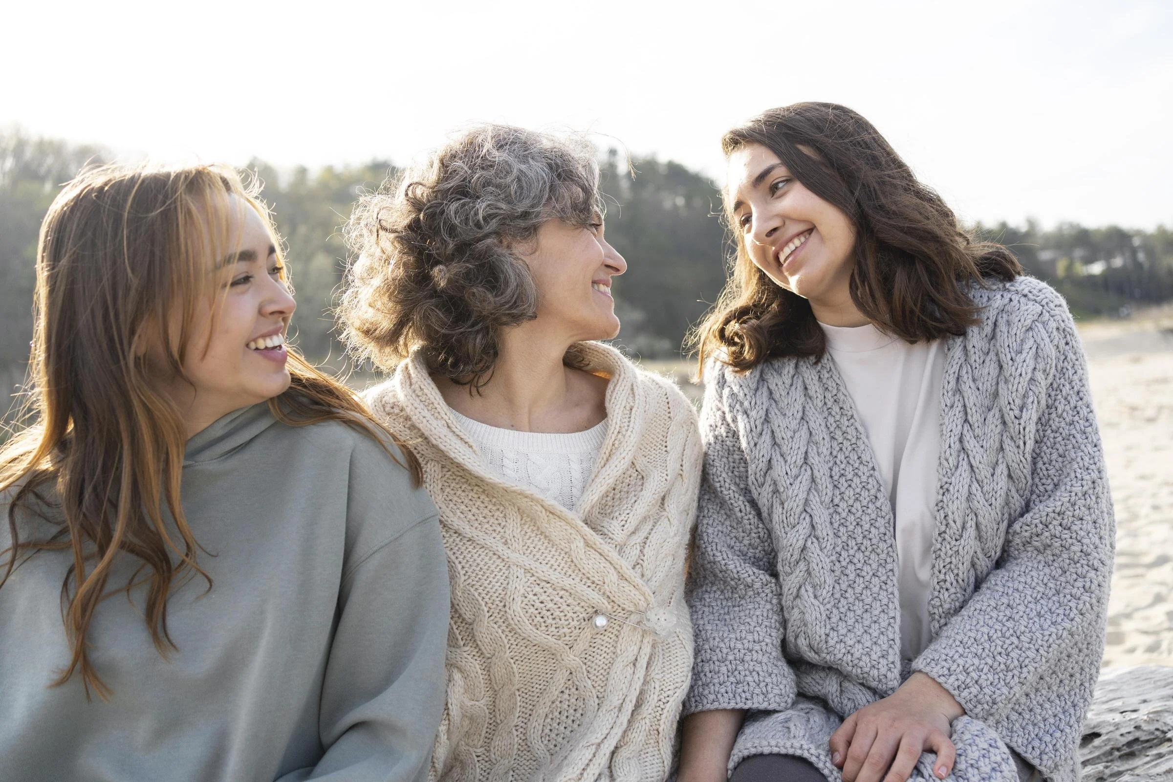 Three women, an elderly woman in the middle and two younger women on either side, smiling and looking at each other outdoors in a natural setting.