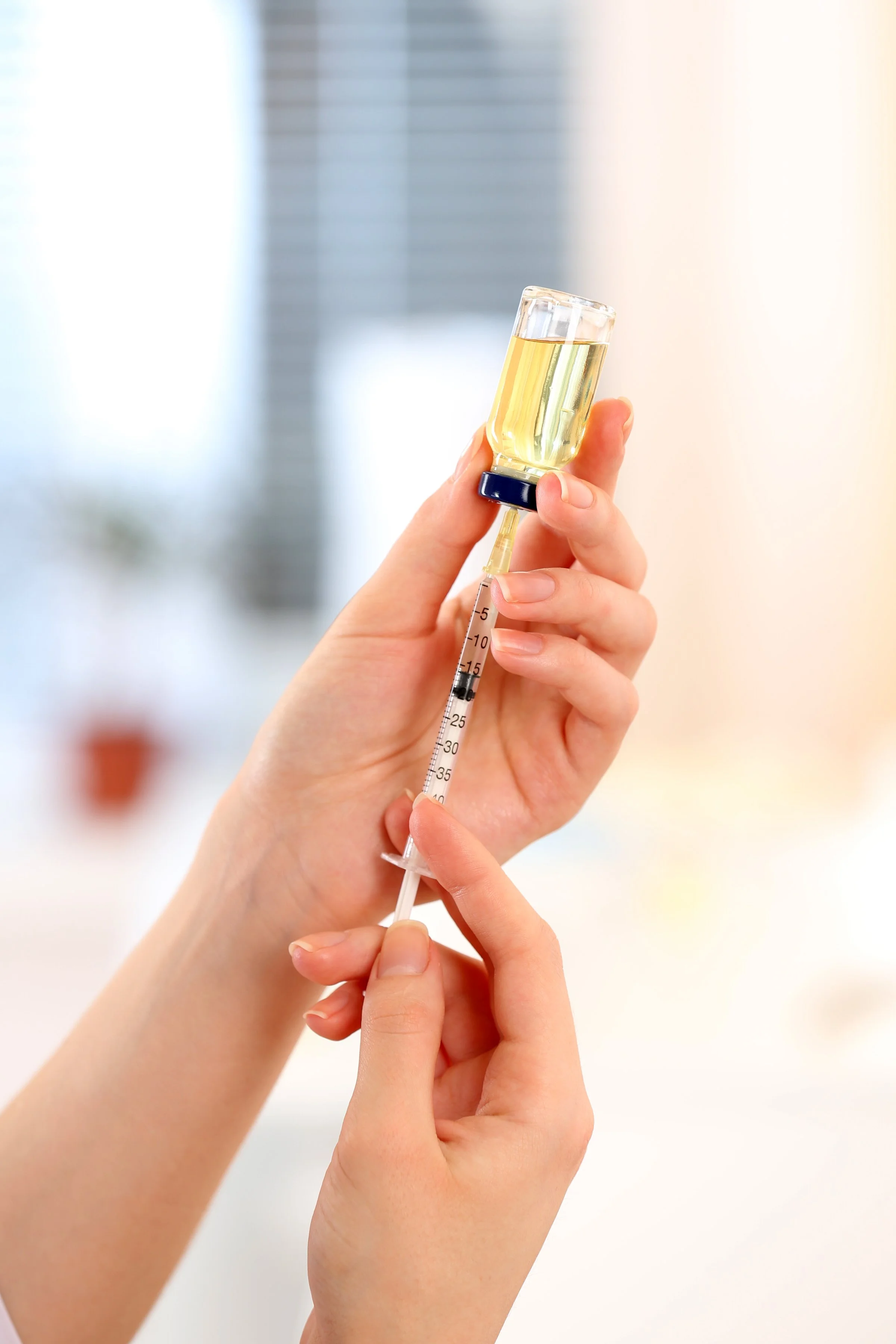 Close-up of a hand holding a syringe with a liquid, in front of a glass vial with a yellowish solution.