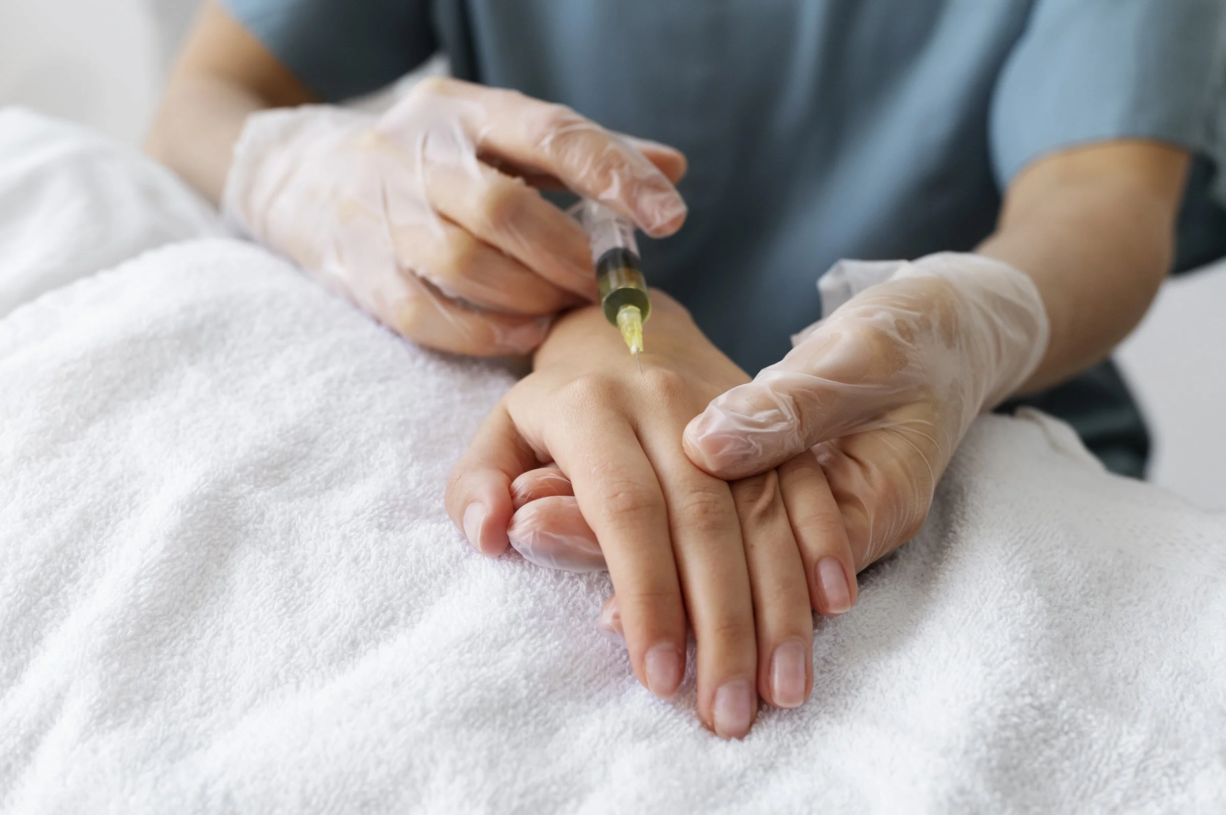 Medical professional administering an injection to a patient’s hand with gloves on, on a white towel surface.