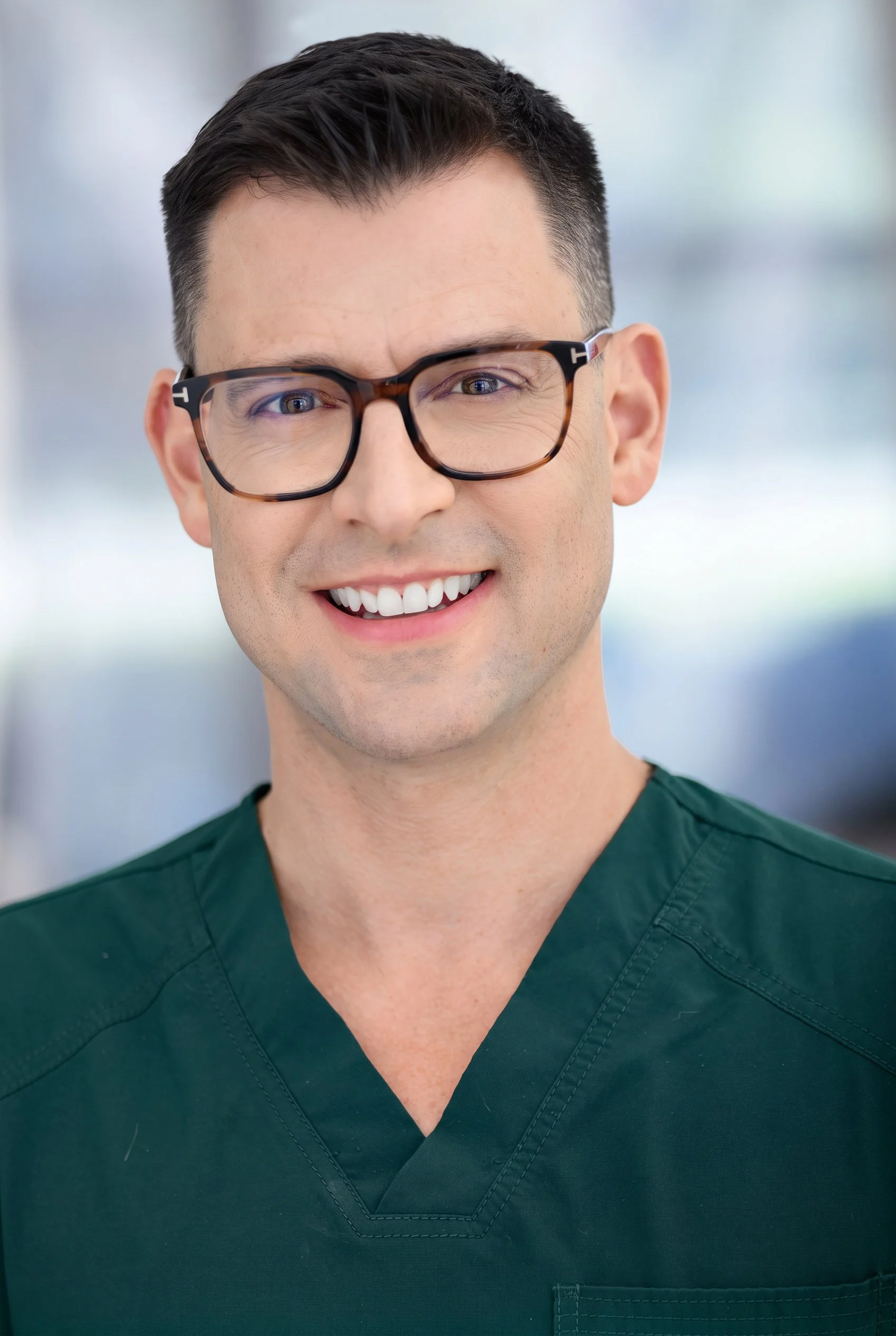 Close-up of a smiling male healthcare professional wearing glasses and green scrubs, with a blurred background.