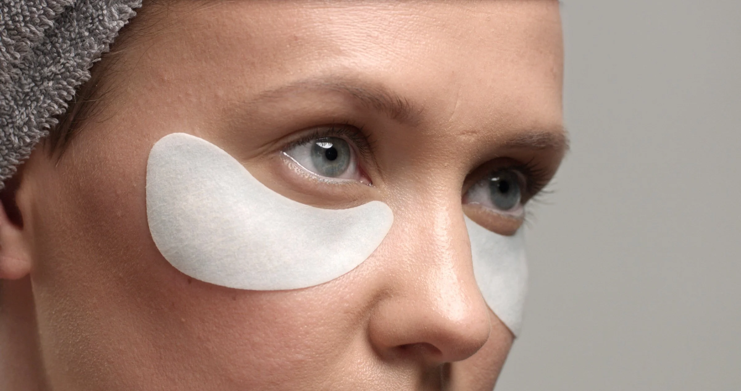 Close-up of woman’s face with a gray towel wrapped around her head and a white under-eye patch on her cheek.