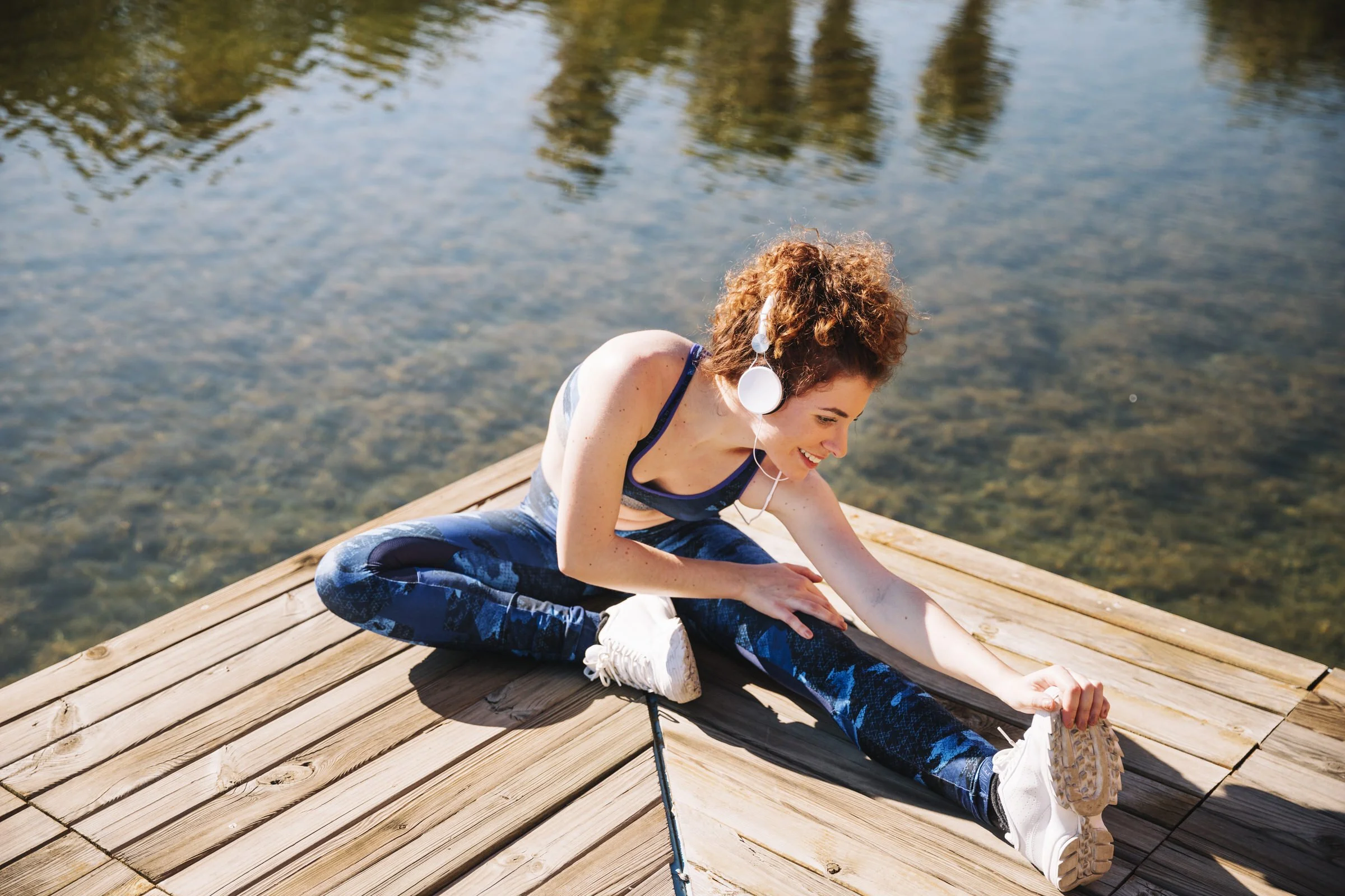 A young woman with curly red hair doing yoga on a wooden dock by a lake, wearing headphones and sportswear.
