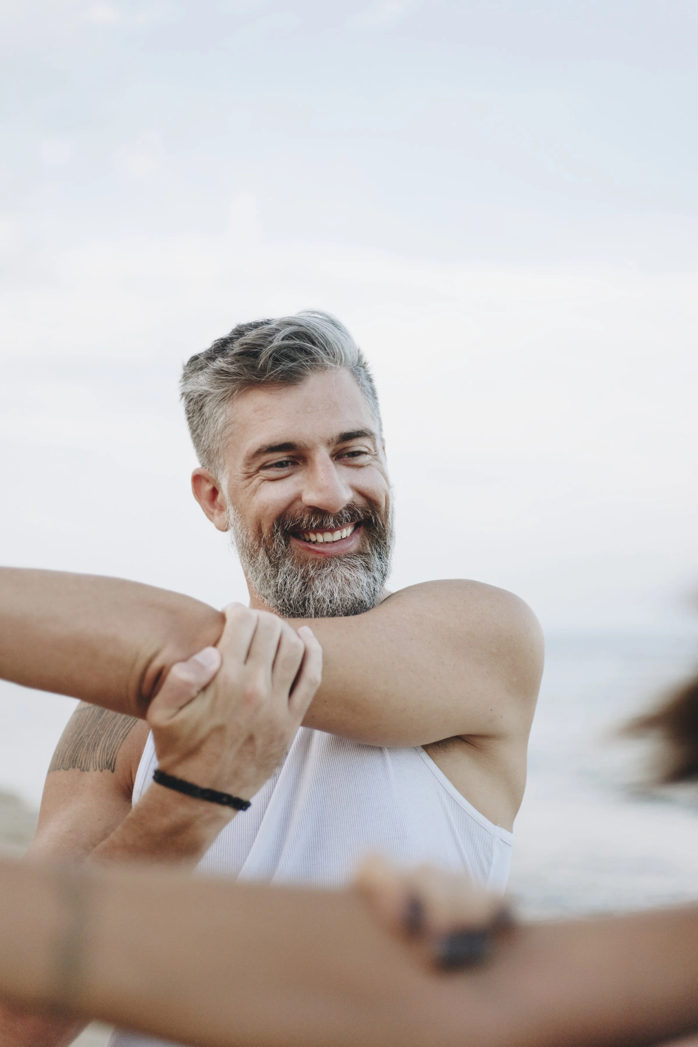 A man with a beard and gray hair smiling, wearing a white tank top, participating in a handshake or embrace outdoors during daylight with a clear sky background.