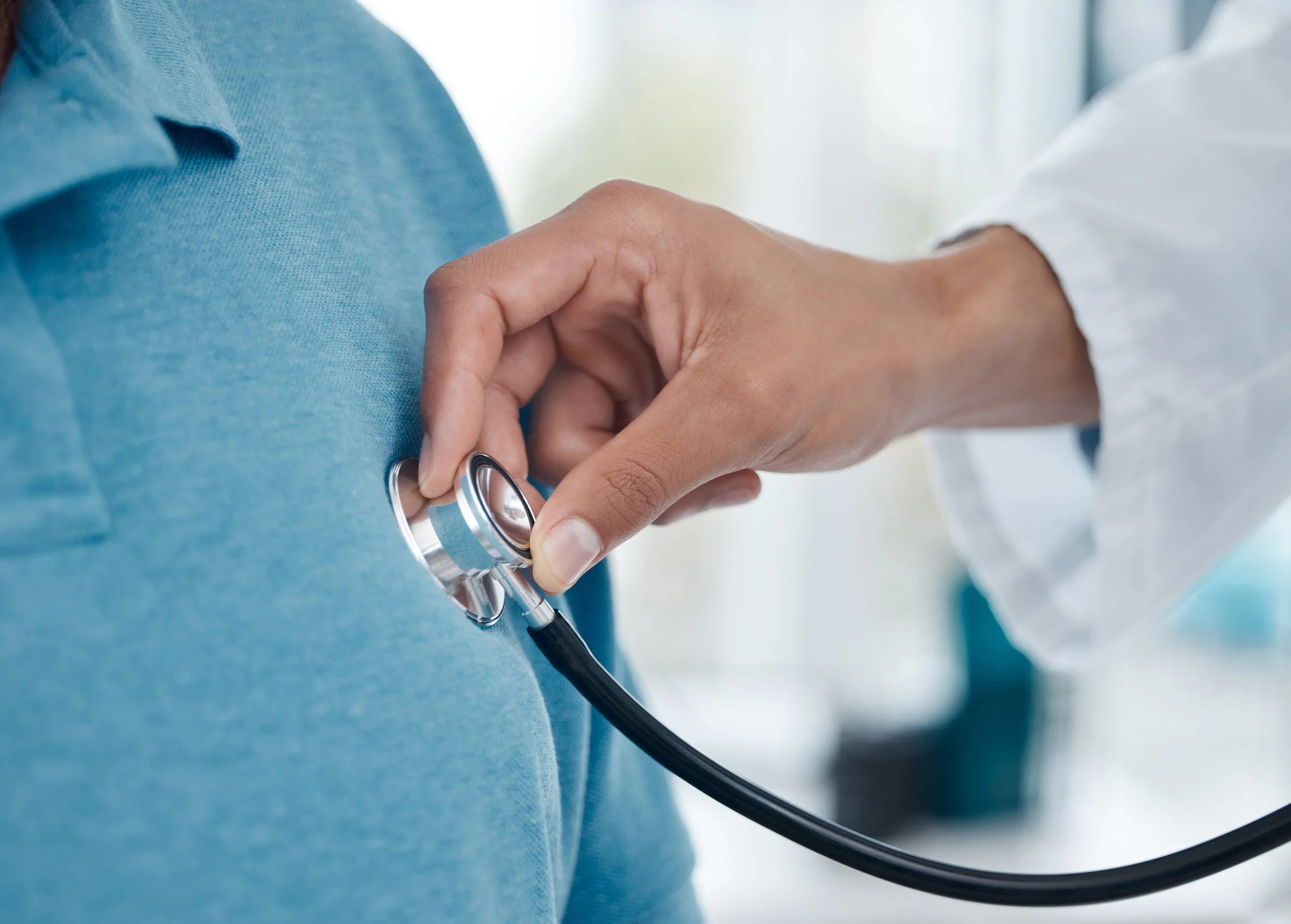 A close-up of an osteopathic physician using a stethoscope on a patient's chest, who is wearing a blue shirt, in a clinical setting with natural light.