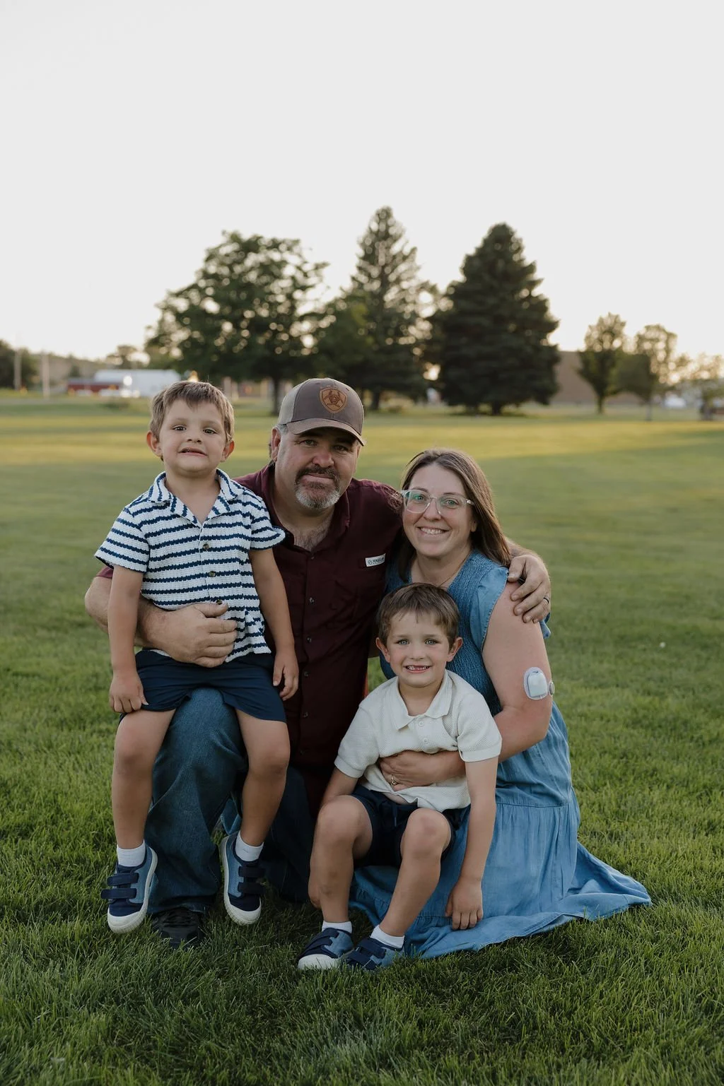 Family of four outdoors on a grassy field with trees in the background, smiling and posing for a photo.